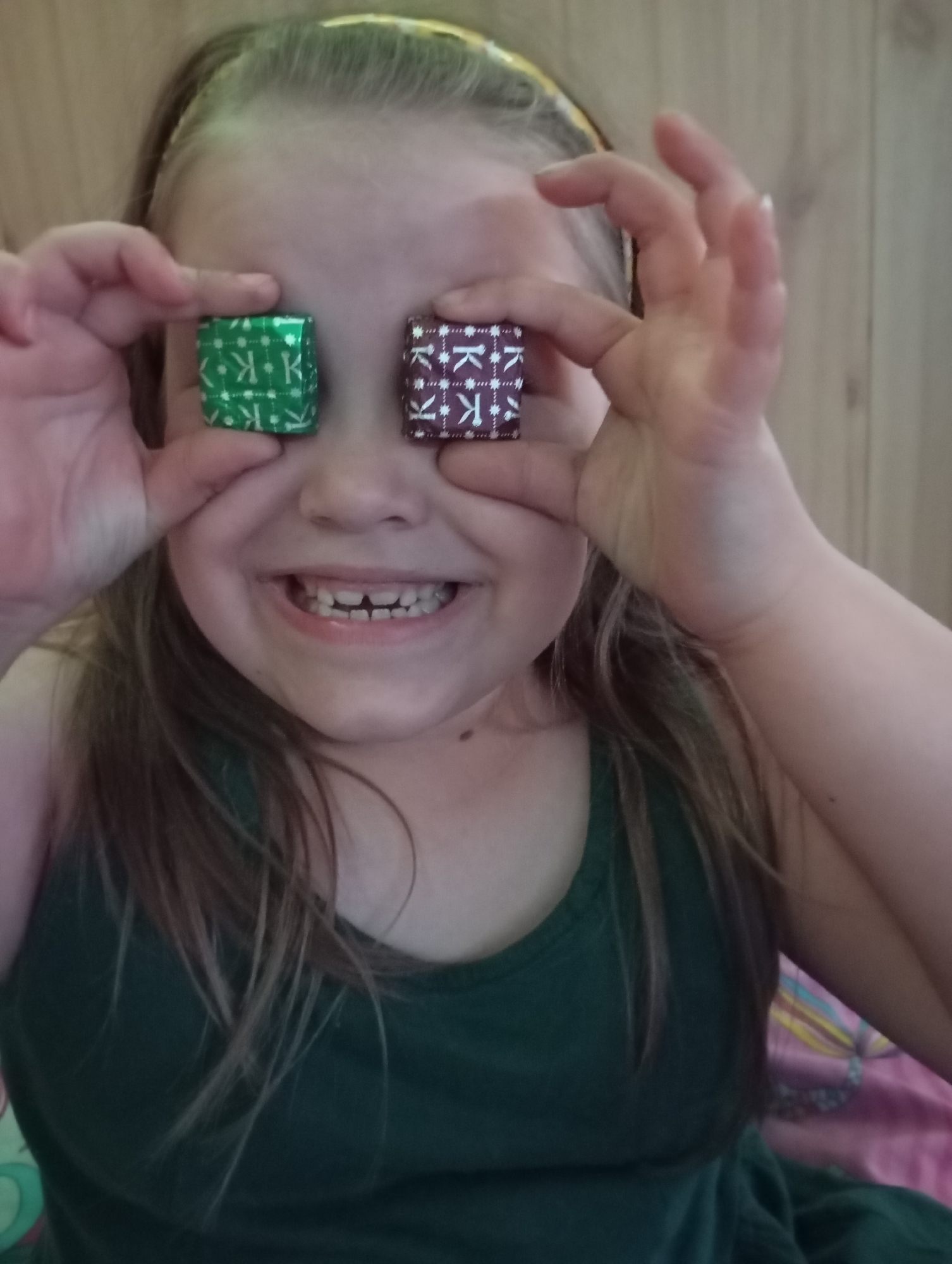 A young girl is holding two wrapped candies in front of her eyes. The candy on the left is wrapped in green foil with white "K" logos, and the candy on the right is wrapped in dark purple foil with white "K" logos. She is smiling broadly, showing her teeth, and wearing a yellow headband. She is wearing a dark green sleeveless shirt. The background appears to be a wooden wall and part of a pink patterned bedspread. The location is Unknown, Unknown.