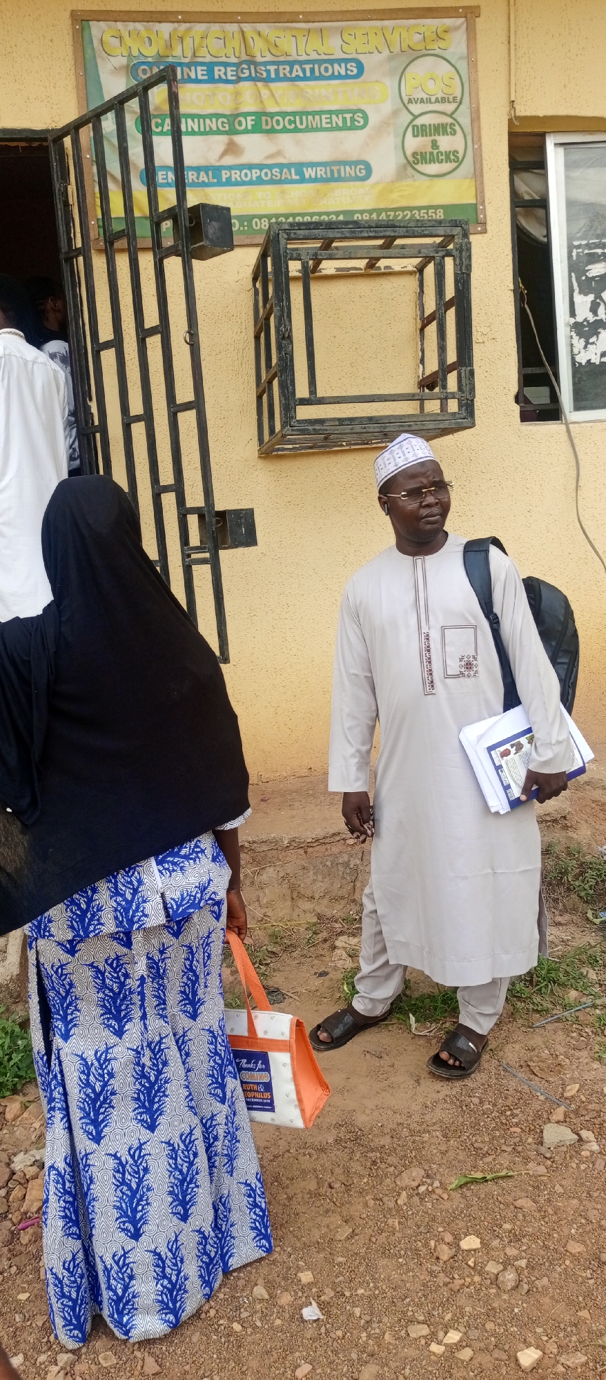 The image depicts an outdoor scene in Keffi, Nigeria, featuring two people standing outside a building. On the left, a person is viewed from behind, dressed in a black hijab and a long blue and white patterned skirt or dress. They hold an orange and white reusable bag that reads "Thanks for COMING RUTH & EOPHILUS." To their right, a man with glasses and a white patterned cap (kufi) wears a light-colored traditional long-sleeved garment (thobe/kaftan) with embroidered details. He has a black backpack on his left shoulder and carries a stack of papers or notebooks in his left hand, looking off to the right.
The building behind them has a textured, yellowish-tan wall, with a black metal gate or window bars visible. Mounted on the wall is a signboard for "CHOLITECH DIGITAL SERVICES." The sign advertises services such as "ONLINE REGISTRATIONS," "PHOTOCOPYING," "SCANNING OF DOCUMENTS," and "GENERAL PROPOSAL WRITING." It also notes "POS AVAILABLE" and lists "DRINKS & SNACKS." Two phone numbers, "08404000034" (partially visible) and "09447223558," are printed at the bottom of the sign. The ground is unpaved, composed of dirt and scattered small rocks. The scene is brightly lit, indicating daytime.