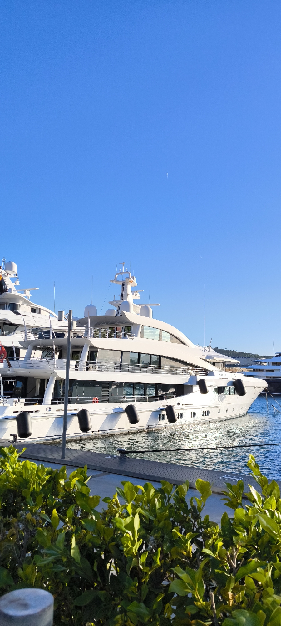 A daytime view of a large, multi-decked, white luxury yacht docked at a pier in Barcelona, Spain. The yacht is positioned to the right of another similar, though slightly smaller, vessel. Both yachts have multiple decks and a series of black fenders along their hulls. The foreground contains green foliage of bushes obscuring the bottom of the pier. The water reflects sunlight, causing a shimmer on the surface. A clear, bright blue sky fills the upper portion of the image.