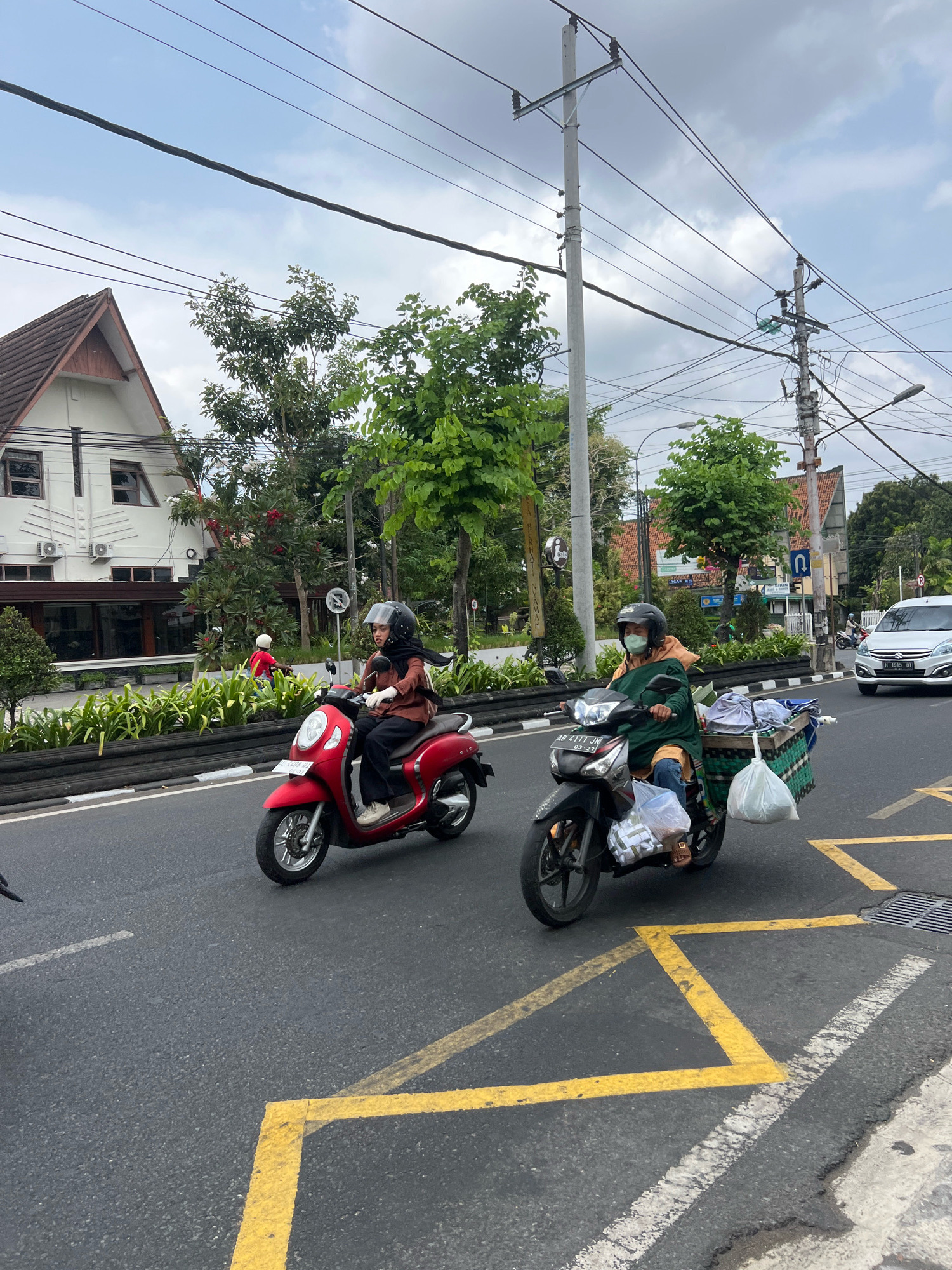 Two people on motorcycles are traveling down a city street in Yogyakarta, Indonesia. The person on the red motorcycle is wearing a brown shirt, black pants, and a black helmet. They are looking to the left. The person on the dark-colored motorcycle is wearing a green robe over a hoodie, blue jeans, and a black helmet with a light green face mask. They are looking straight ahead. The dark-colored motorcycle has a green basket attached to the back, filled with items. The street has yellow lane markings. In the background, there are buildings, trees, power lines, and a white car. The sky is cloudy.
