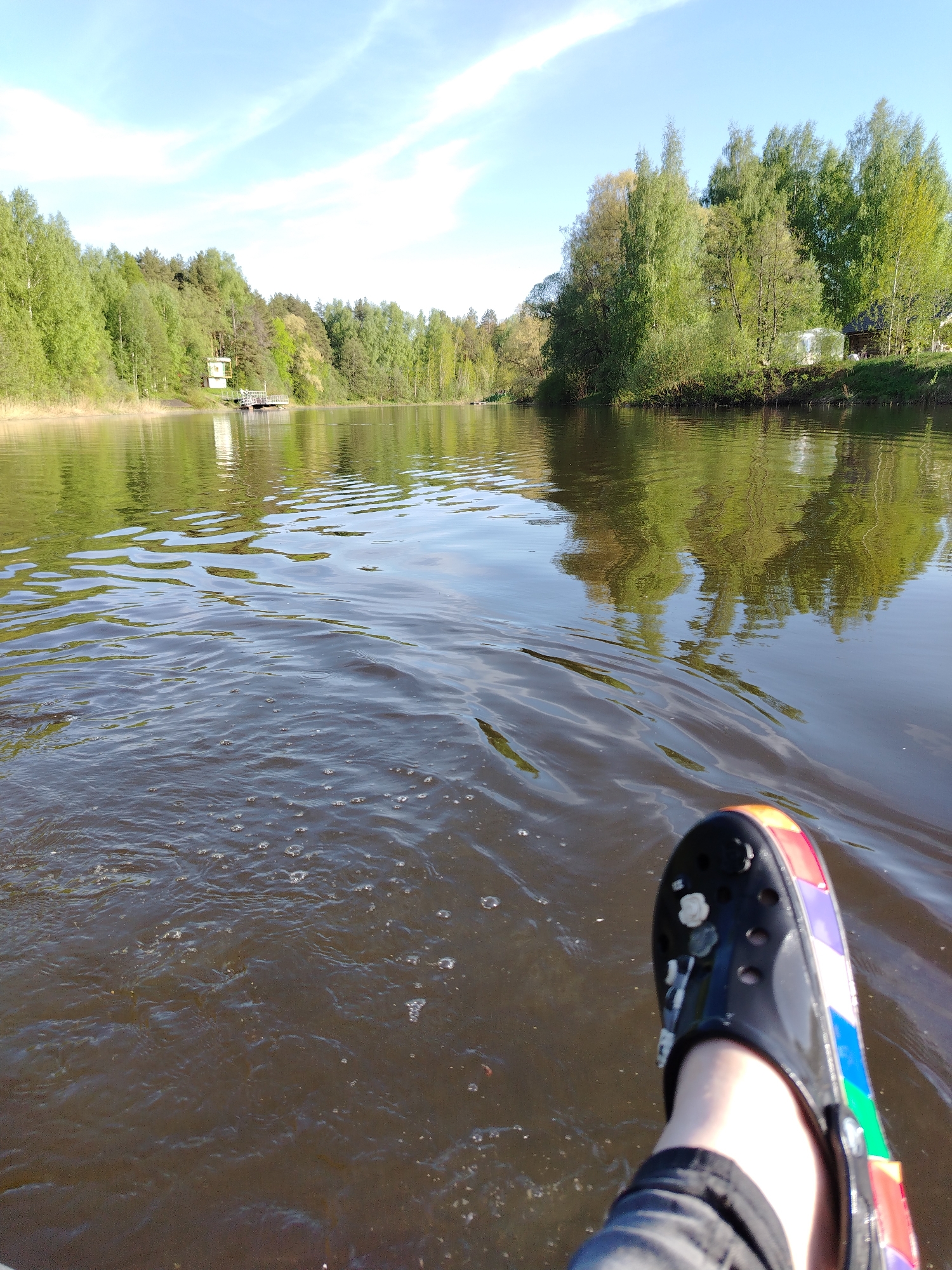 A point-of-view shot depicts a person seated in a small watercraft on a body of water, likely a river or lake, in Stroitel', Russia. The person's left leg, clad in gray pants and wearing a black Croc shoe with a rainbow-colored trim, extends into the foreground. The water, rippled by the motion of the boat, is dark and reflective, mirroring the dense green foliage of trees lining both banks. In the distance, structures are visible along the shoreline, suggesting a populated area. The sky is light blue with wisps of clouds. The scene appears to be taken on a bright, sunny day.