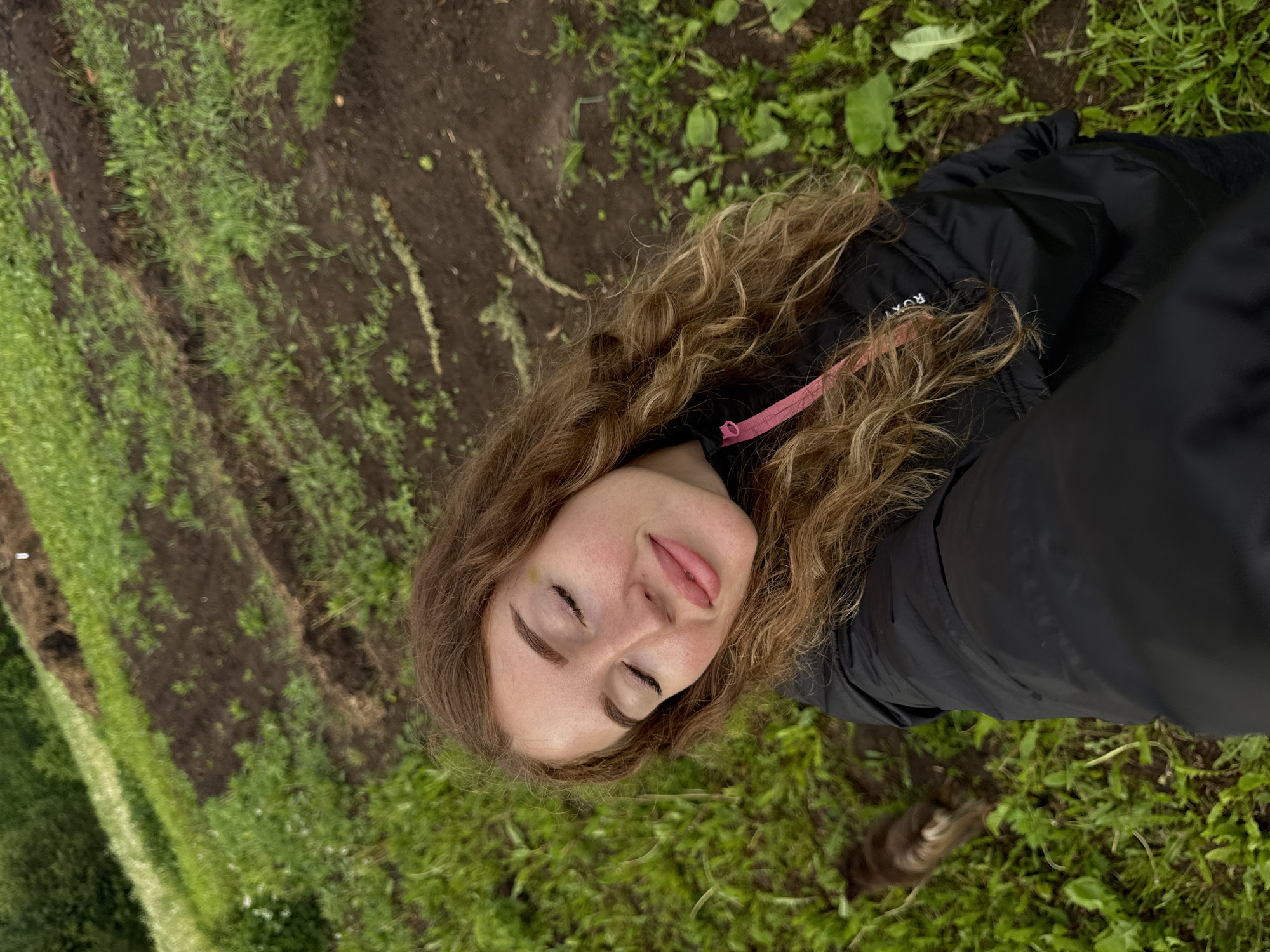 The image captures a young woman in an outdoor setting, identified as Maksimovo, Russia. She has long, curly, light brown hair and appears to be looking upwards with her eyes closed and a serene expression, her lips slightly parted. A faint smudge is visible on her right cheek. She is wearing a black puffer jacket with a pink zipper pull, and the brand name "ROXY" is partially visible on the jacket's chest.
The photograph is taken from an overhead perspective and is rotated, showing the ground vertically. The background is a natural landscape composed of patches of vibrant green grass interspersed with areas of dark brown soil, suggesting a field, garden, or natural path. The ground appears somewhat damp, implying recent moisture. The lighting is soft and diffused, indicating daytime, possibly under an overcast sky, as there are no harsh shadows. The overall mood conveyed is one of peace and relaxation.