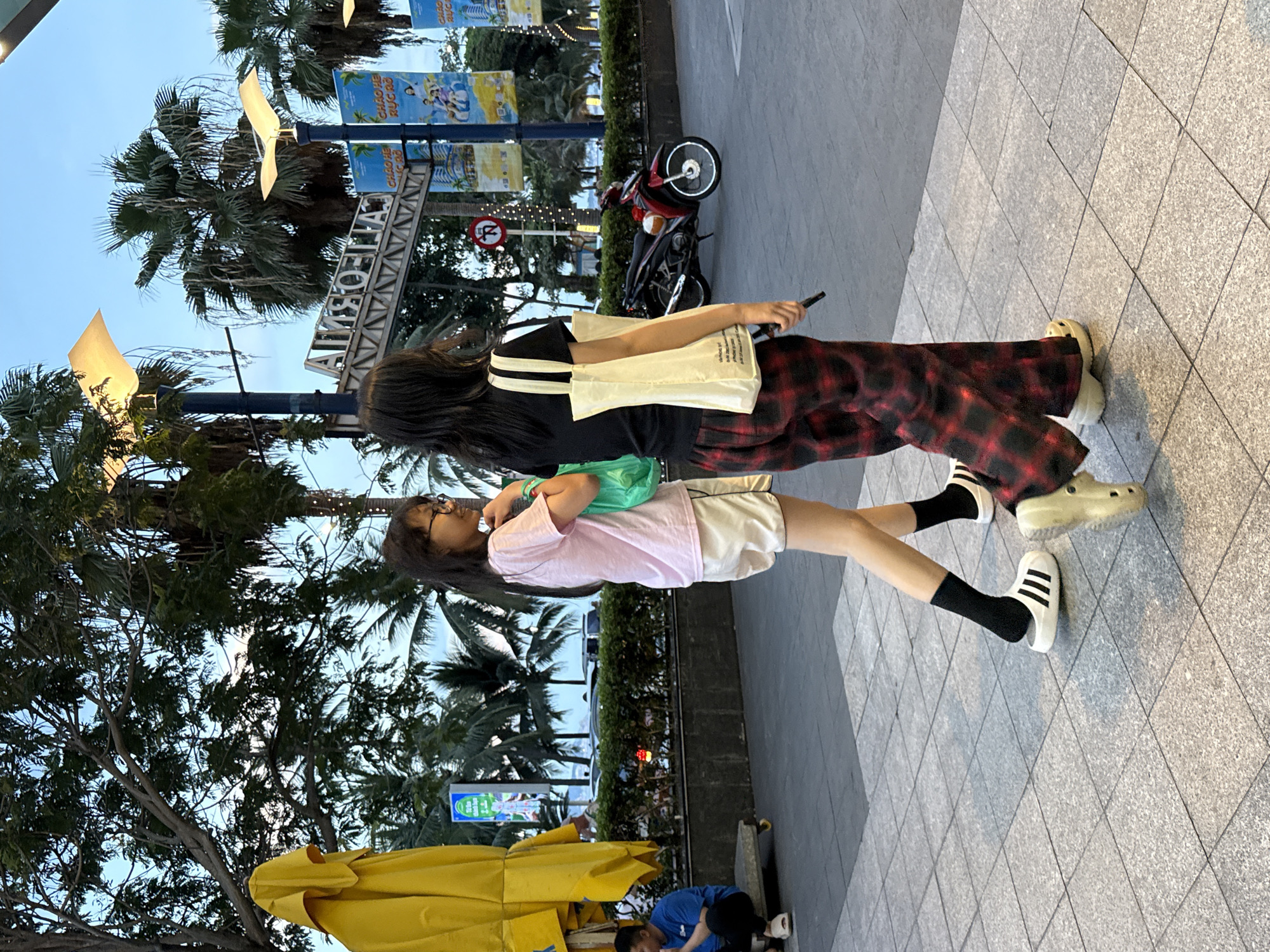 In Nha Trang Ward, Vietnam, two young women are walking on a tiled pedestrian path. The woman on the left wears a pink t-shirt, white shorts, black socks, and white Adidas slides. She carries a green bag in her right hand. The woman on the right wears a black t-shirt, red and black plaid pants, and light-colored Crocs. She carries a beige tote bag in her left hand and holds a black object in her right hand. Behind them, palm trees and a blue sky are visible. A red and black motorcycle is parked to the right, and a sign with Vietnamese text and images of people is displayed on a pole. A yellow tarp covers an object to the left. A person in a blue shirt is seated on a small cart in the background.