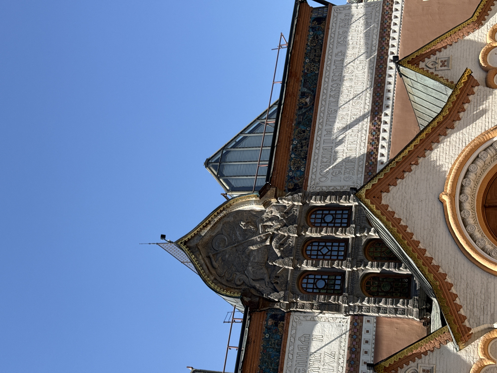 The image shows a section of the State Historical Museum in Moscow, Russia, tilted with a clear blue sky in the background. The museum's facade is richly decorated with intricate carvings, inscriptions in Cyrillic script, and small windows. There's a mix of architectural styles, including decorative brickwork, stone carvings, and a small section with glass panels. The angle suggests a view from ground level, looking upwards at a portion of the building's detailed exterior.