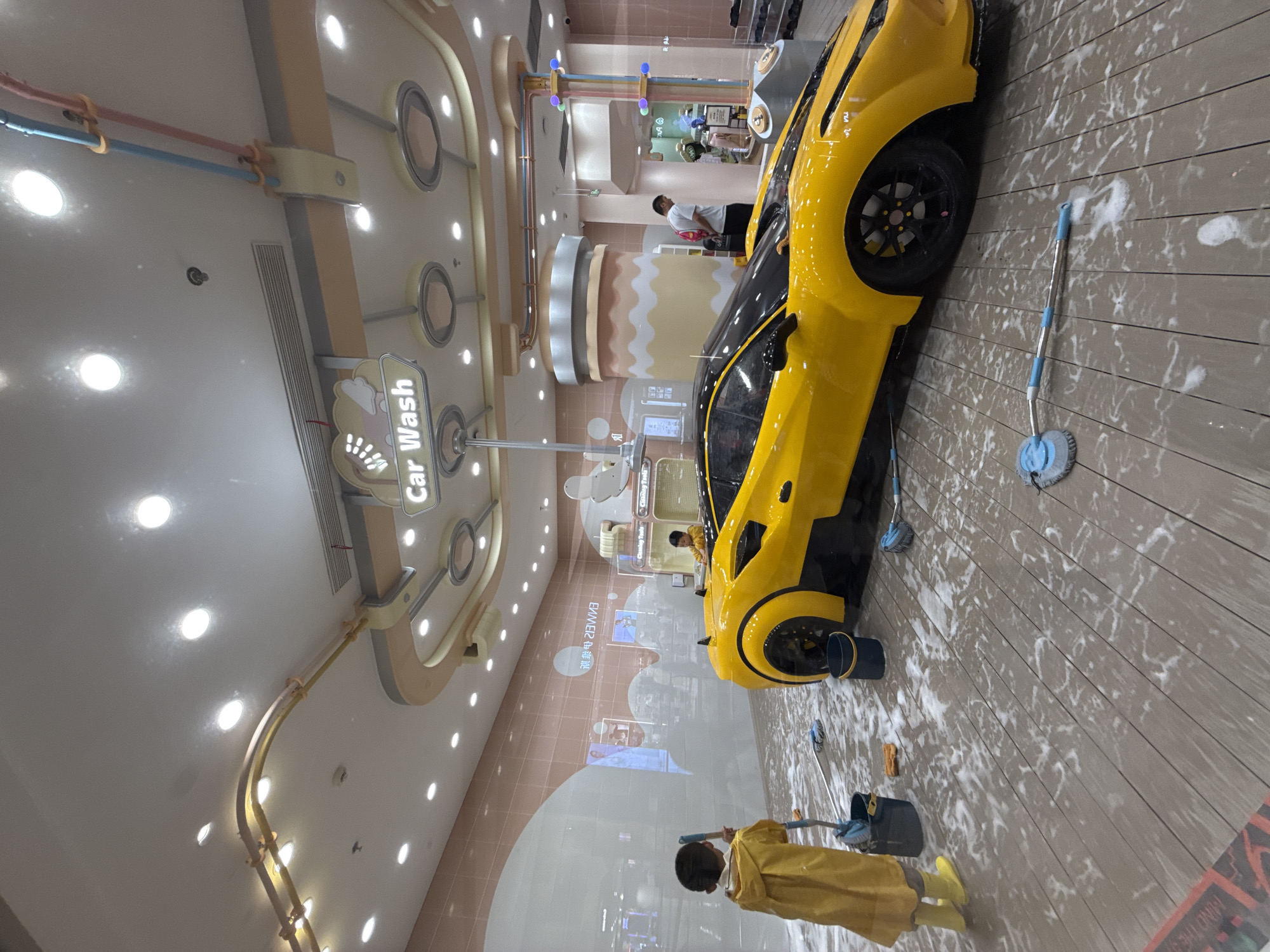 The image depicts a children's car wash play area within a commercial establishment. The scene is set on a light brown, wooden-planked floor, which is visibly wet and covered in white soap suds. In the center, a bright yellow sports car, possibly a replica, is the main attraction. A young child, dressed in a yellow raincoat and yellow boots, stands to the left, holding a blue-handled mop with a brush head, actively engaged in cleaning the floor around the car. Nearby, a dark blue bucket sits on the floor. Another child, wearing a yellow shirt, is visible in the background to the right, near the car, appearing to interact with it. The background features a pink-tiled wall with decorative elements, including a prominent "Car Wash" sign with cartoonish imagery, suspended from the ceiling. Above, the ceiling is illuminated by numerous circular recessed lights. The overall environment suggests an indoor recreational space designed for children's imaginative play, located in Liangjiang, China. The presence of the car, cleaning tools, and suds indicates a simulated car washing activity.