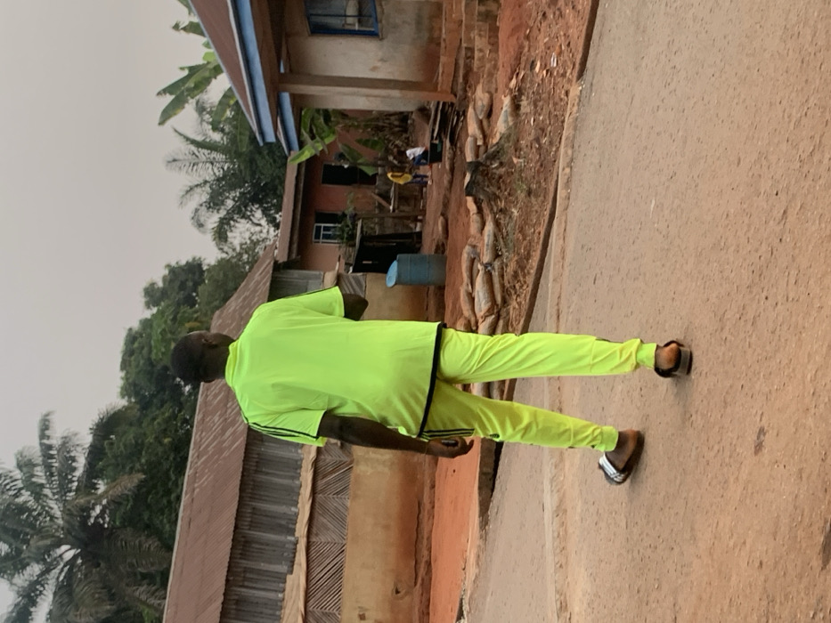 A man in a bright yellow tracksuit stands on a road in front of some buildings. The buildings appear simple, with one showing signs of wear. A pile of firewood or logs rests beside one of the buildings. The sky is overcast, and the environment suggests a rural or less developed area. Palm trees are visible in the background. The man is wearing sandals.