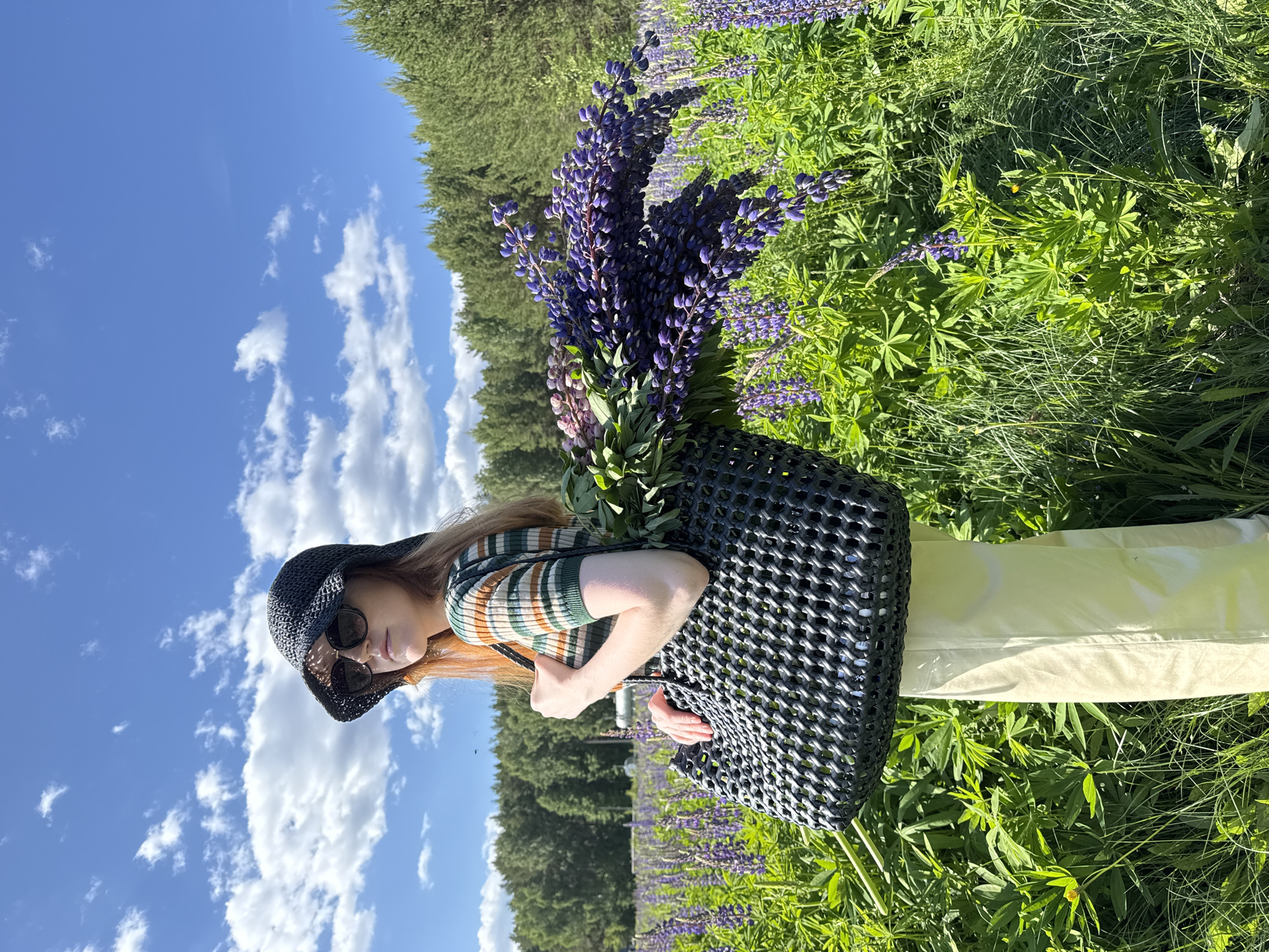 A female individual stands outdoors in a field identified as being in Chaikovsky, Russia, under a clear blue sky with scattered white clouds. The individual, with reddish-brown hair, wears sunglasses and a dark woven bucket hat. She is dressed in a horizontally striped short-sleeved top featuring green, brown, and off-white bands, paired with light-colored (possibly cream or beige) wide-leg trousers. She carries a large, dark-colored, possibly black, woven tote bag over her right shoulder, which is filled with freshly gathered purple lupin flowers and their green foliage, overflowing from the top. The surrounding environment consists of a dense field of blooming purple lupins and other green vegetation. In the background, a line of dense green trees forms a distant forest edge. The scene is brightly lit by natural sunlight.