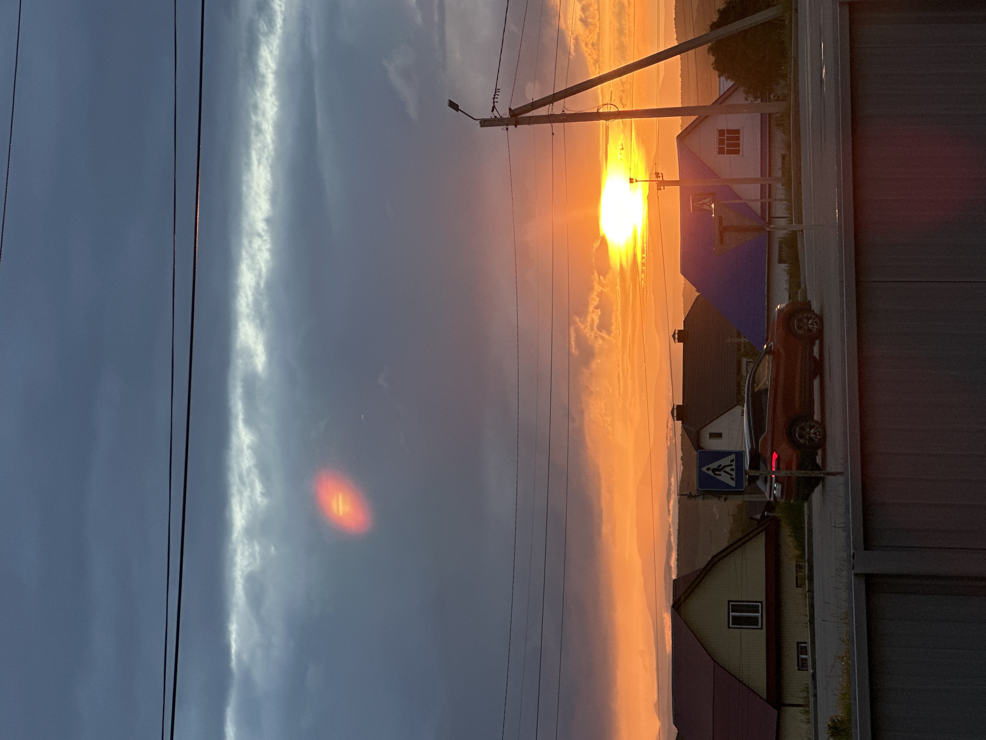 The image captures an outdoor scene at either sunrise or sunset, characterized by dramatic lighting and a residential setting. The sky is a prominent feature, transitioning from brilliant orange and yellow hues near the horizon where a dazzling sun is visible, to a darker blue-grey on the left. Long, wispy clouds stretch across the sky, with one distinct, bright, elongated cloud formation running vertically in the darker section. A red, elliptical lens flare with horizontal striations is also present in the upper left sky.
Utility poles with numerous power lines traverse the upper half of the frame. One pole includes a street light. Below the horizon line, several houses are visible. One prominent house has a white facade with a red-brown roof and a white-framed window. Another building features a distinctive blue roof. In the foreground, a paved road or driveway runs along the right side, bordered by a dark, corrugated fence or wall. A red SUV-like vehicle is parked or positioned on the road. Adjacent to the road, a blue triangular road sign with a white border displays a white pedestrian crossing symbol.
No people are visible, and no specific activity is overtly taking place, giving the scene a tranquil, static quality. The location appears to be a suburban or rural residential area, possibly alongside a main road. There is no discernible text visible in the image.