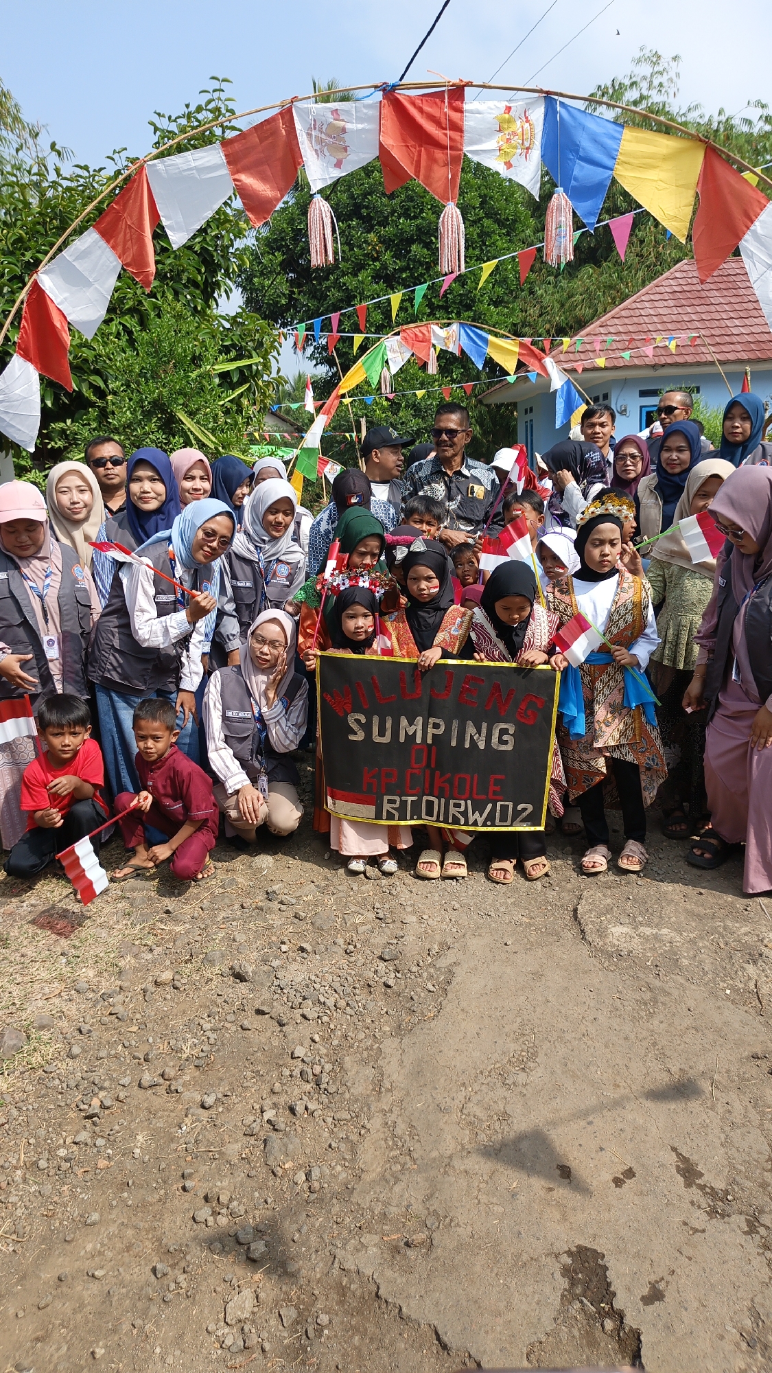 In Linggasirna, Indonesia, a group of people, predominantly children, are gathered outdoors under a festive arch adorned with red and white flags and colorful pennants. The ground is unpaved, composed of dirt and small stones, with a path visible in the foreground. The group appears to be posing for a photograph, with many holding small Indonesian flags. A large black banner with white lettering is held by several children, reading "WILUJENG SUMPING DI KP. CIKOLE RT. OIRW.02". Behind them, trees and a residential building with a red tiled roof are visible. The atmosphere seems celebratory, possibly for a local event or holiday, indicated by the decorations and the presence of both children and adults.