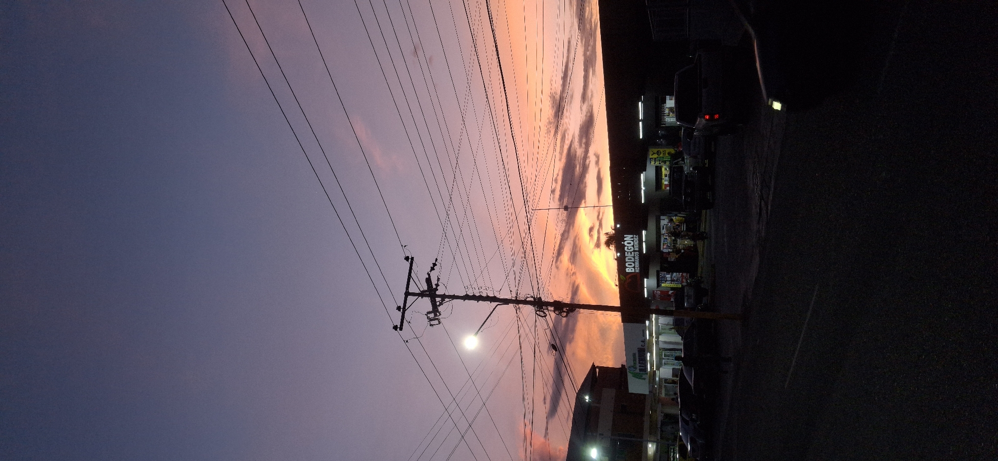 The image depicts a street scene in Tinaquillo, Venezuela at dusk. An electric pole with a streetlight stands on the right side of the road, with many electrical wires extending across the sky. The sky fades from dark blue to orange-pink as the sun sets. A "Bodegón Hermanos Méndez" store is situated on the right side of the image. A few cars are parked on the street. The road surface appears dark, suggesting asphalt.