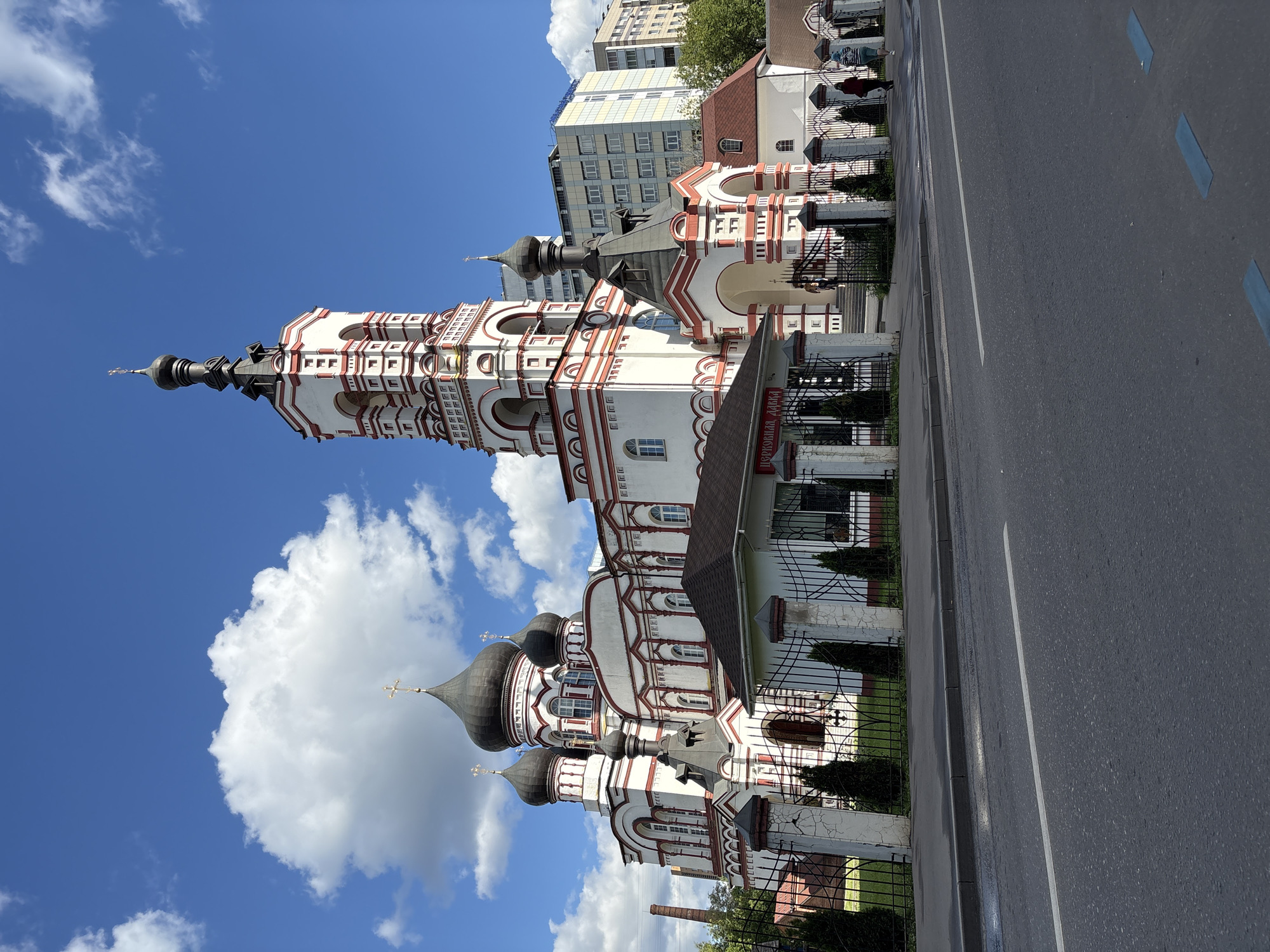 The image shows a white Orthodox church with red trim and multiple domes, topped with golden crosses, set against a bright blue sky with scattered white clouds. A tall, slender bell tower with red and white banding stands to the right of the main church structure. In front of the church, a low-rise building with a dark roof and visible windows is situated behind a black metal fence and stone pillars. The church complex is bordered by a fence with ornate metalwork, and manicured evergreen trees are planted along the fence line. A paved road runs in the foreground, with a white stripe marking a lane. Several people are visible near the entrance of the church and on the sidewalk. In the background, modern multi-story buildings are visible. The overall scene suggests a sunny day in an urban environment. The church is located on the intersection of Tverskaya-Yamskaya Street and Sadovaya-Karetnaya Street in Moscow, Russia.