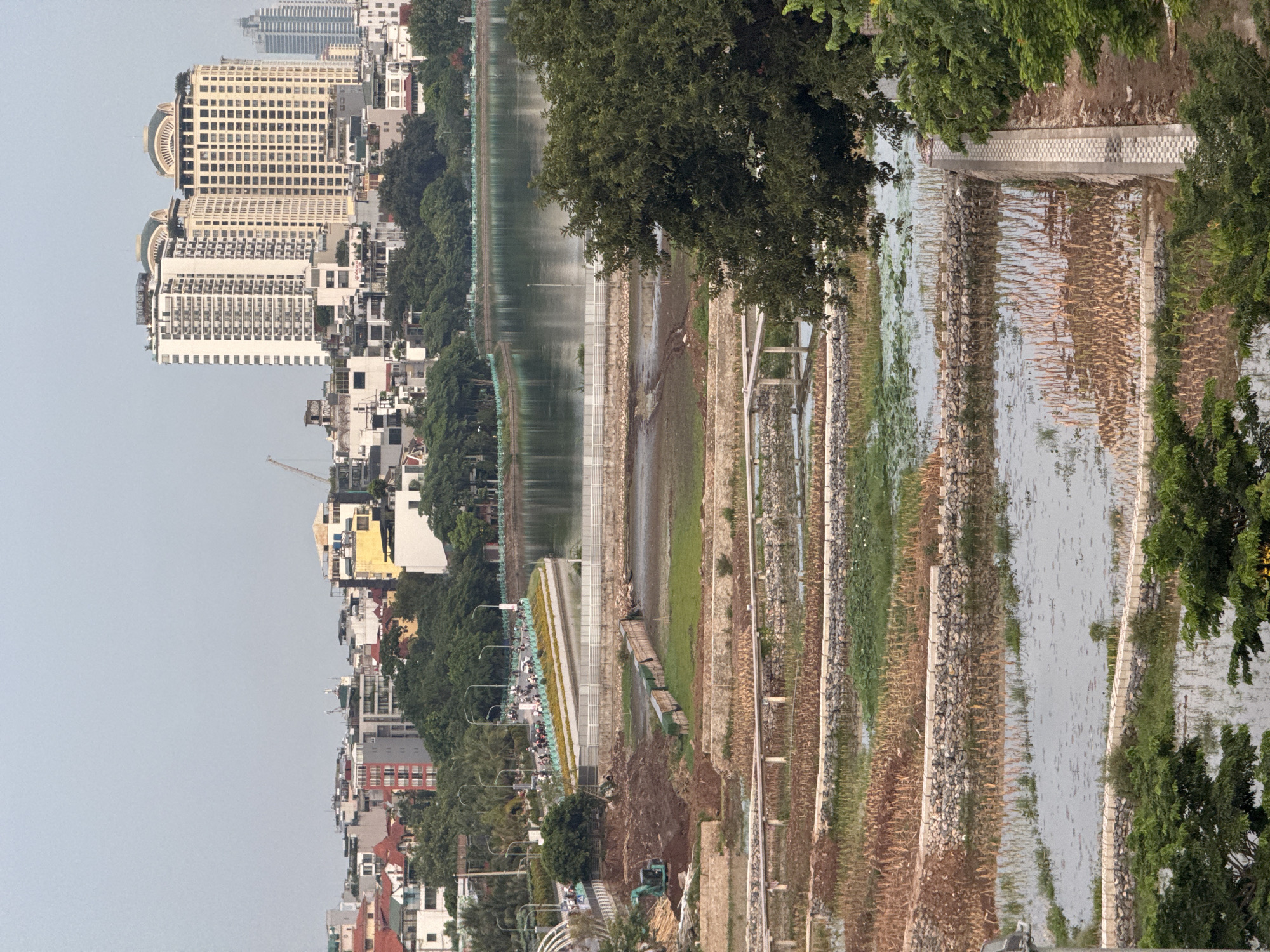 The image depicts a waterfront area in Hanoi, Vietnam. The scene is dominated by a body of water on the right, with a tiered embankment on the left that appears to be under construction or recently renovated. The embankment is composed of layers of stone and vegetation, with some areas showing exposed soil and construction equipment, including an excavator. Beyond the embankment, there is a road with a line of trees and a pathway bordered by a white fence. Further to the left, a dense urban landscape rises, featuring a variety of buildings, including tall, modern apartment buildings and older, lower-rise structures. Trees are interspersed throughout the urban area, and a distant skyscraper is visible. The water's surface is rippled, suggesting a gentle breeze or light rain. The sky is overcast, indicating a cloudy day, possibly in the early morning or late afternoon due to the soft light. The overall impression is one of a developing urban environment alongside a natural or man-made water feature.