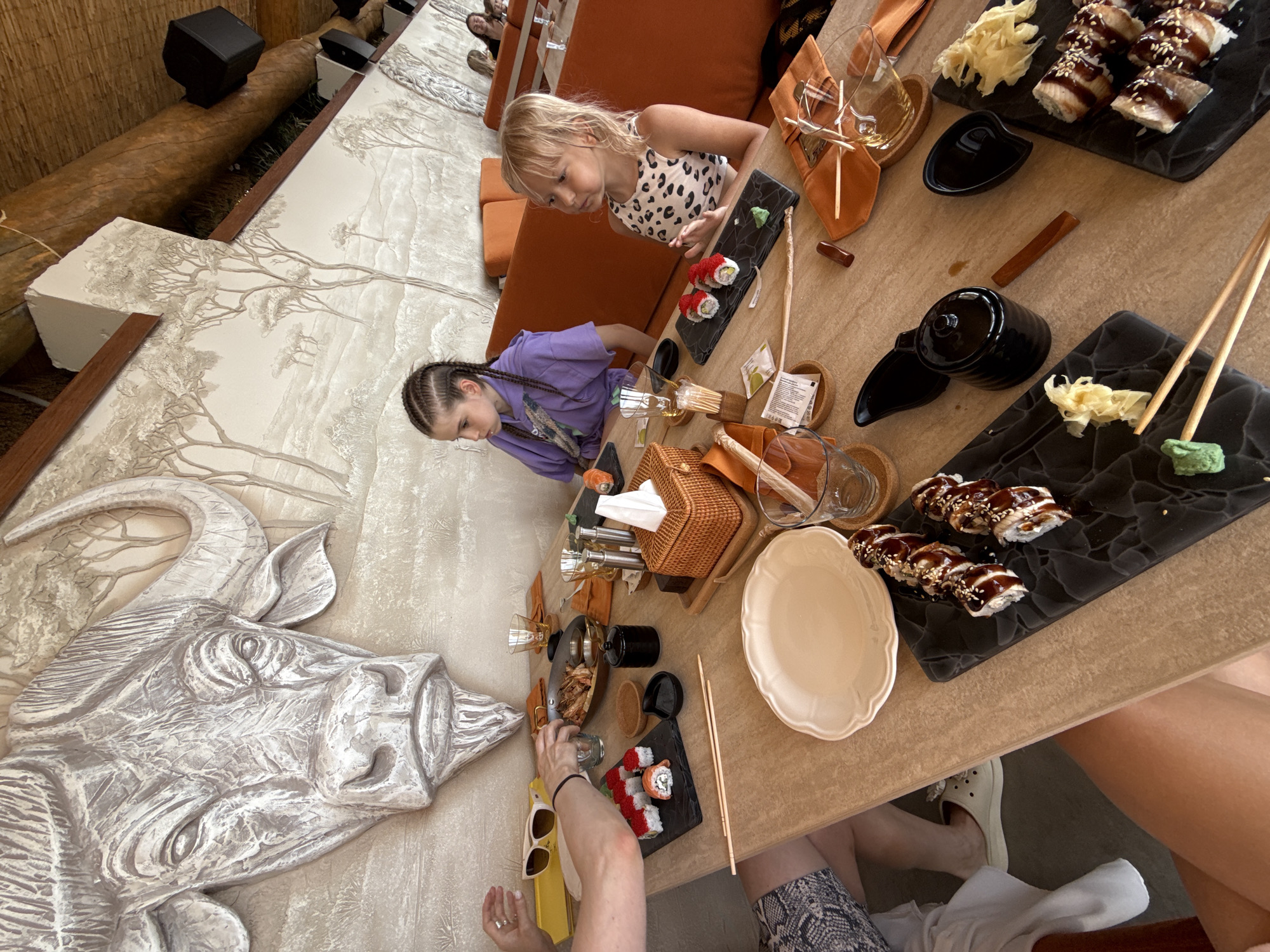 The image depicts a dining scene inside a restaurant, possibly a sushi restaurant, with an African wildlife-themed mural adorning the wall behind the diners. Two young girls are seated at a table, with sushi and drinks laid out before them. The girl on the left has her hair styled in braids, while the girl on the right has blonde hair. Other people are partially visible at the table, suggesting a family or group outing. The table is set with plates, chopsticks, soy sauce dishes, and glasses. The overall ambiance appears to be casual and relaxed. The location is specified as Alushta, Ukraine, though the image itself does not contain explicit visual cues confirming this location.