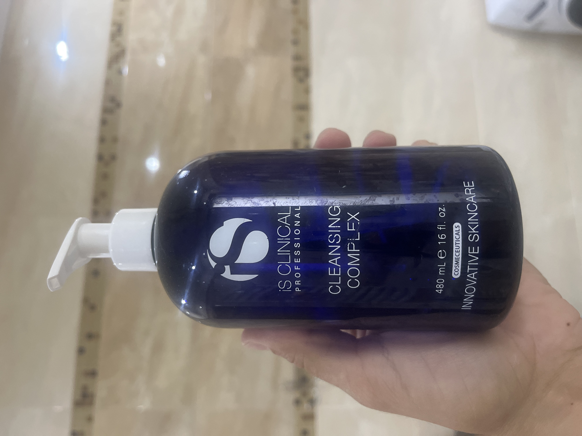 A hand holds a dark blue pump bottle of IS CLINICAL Cleansing Complex. The bottle has white text indicating the brand, product name, volume (480 mL / 16 fl. oz.), and the words "COSMECEUTICALS" and "INNOVATIVE SKINCARE." The background appears to be a tiled wall. There is no indication of the city of Onokivtsi, Ukraine.