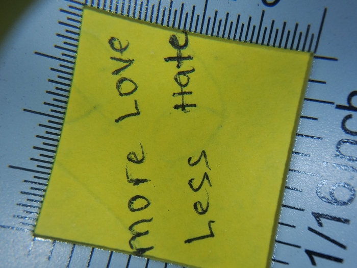 A yellow, approximately 1.5-inch square piece of paper is centrally positioned, resting on a light blue or silver-grey ruler. The paper displays two lines of handwritten text in black ink: "more Love" on the upper line and "Less hate" on the lower line. The underlying ruler features black measurement markings, including numerical fractions like "1/16" and the unit "inch," indicating imperial measurements. Minor surface glare is present on the ruler. The scene is static and devoid of any individuals, actions, or direct interactions. The environment is an unidentifiable indoor setting in Unknown, Unknown.