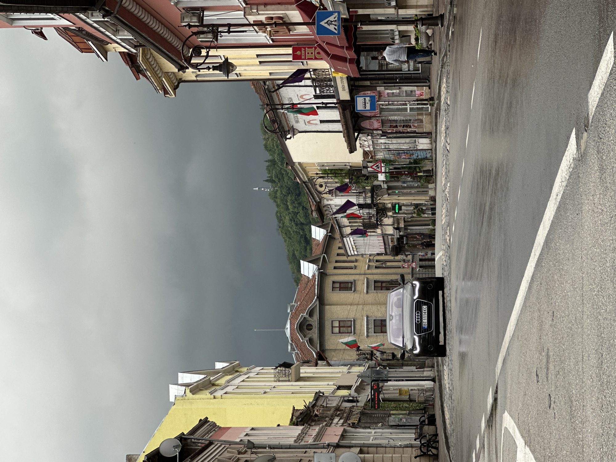 The image shows a street in Veliko Tarnovo, Bulgaria, under an overcast sky. Buildings line both sides of the street. On the left side of the street, buildings are painted in shades of yellow and pink, with architectural details such as eaves and balconies. On the right side, buildings appear to be cream-colored and white. National flags of Bulgaria are visible, hanging from some buildings. A black Audi is parked on the right side of the street, facing the camera. The street surface is wet, reflecting the surroundings. A man is walking along the sidewalk on the right. A forested hill is visible in the distance, framed by the buildings. Street signs are posted along the street.