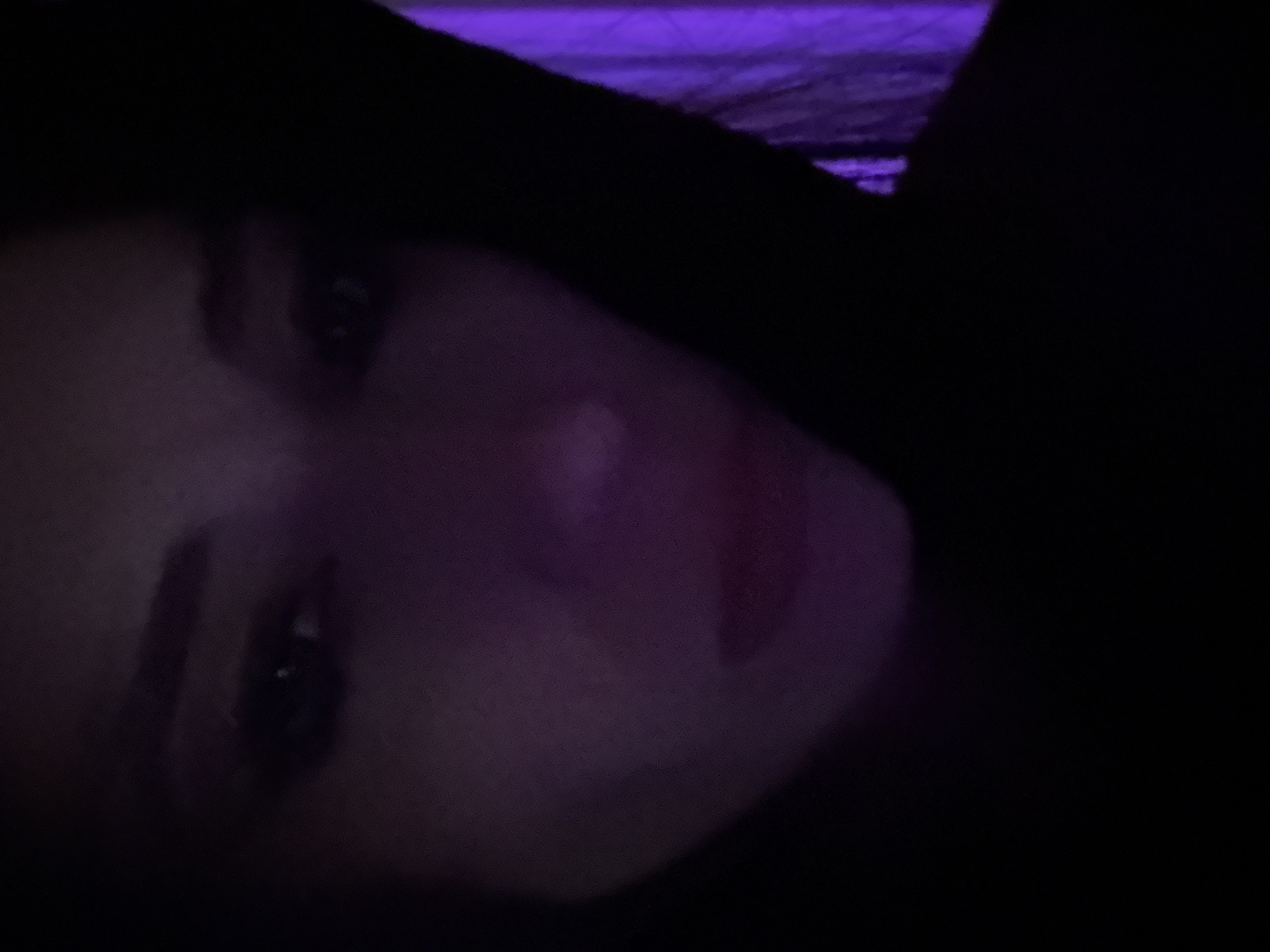 A person's face is partially illuminated with a purplish hue against a predominantly dark background. A hint of purple light is visible in the upper section of the frame. It is hard to discern other details due to the darkness of the image. The location is not discernible from the image.