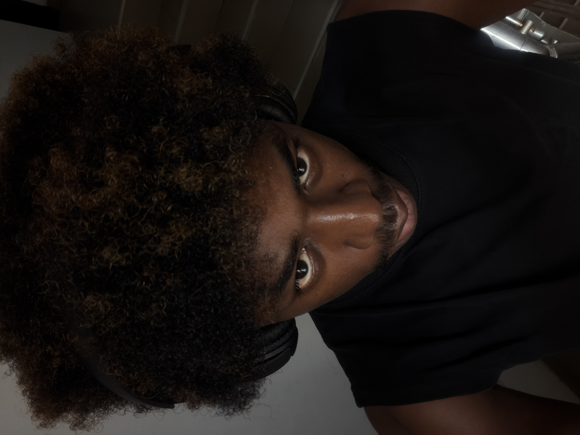 A close-up, overhead shot shows a young Black male wearing a black tank top and black headphones. His dark, curly hair is voluminous and has streaks of lighter brown. His eyes are looking directly into the camera. He has a mustache and goatee. The background is indistinct, with what appears to be a neutral wall on the left and a set of stairs on the right, suggesting an indoor setting. The lighting is dim, with the subject's face illuminated from above. The image does not provide enough detail to determine the specific location within Igboefon, Nigeria.