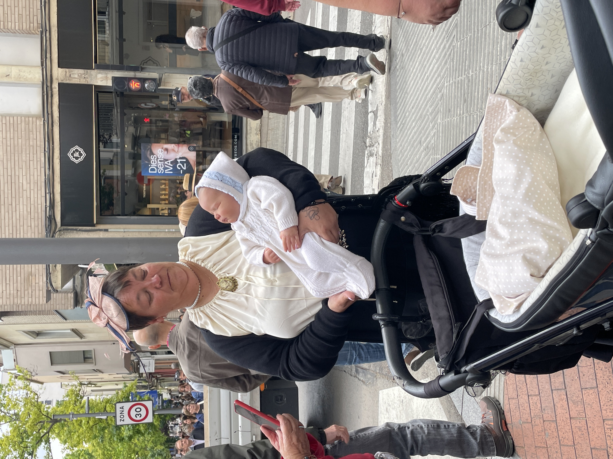 The image captures a street scene in Terrassa, Spain, likely taken during a public event or gathering. The primary subjects are a woman holding a baby, who appears to be sleeping, next to a baby stroller. The woman is wearing a hat with a bow and a dress with a large brooch, and a string of pearls. The baby is dressed in a white knitted outfit with a bonnet. They are situated on a sidewalk with a crosswalk and other pedestrians visible in the background.
Behind them, buildings with shops can be seen, including one with a display advertisement. There is a street sign that shows the speed limit "30". The surrounding environment includes street lights, trees, and a crowd of people in the distance. The overall impression is a moment captured during a daytime event.