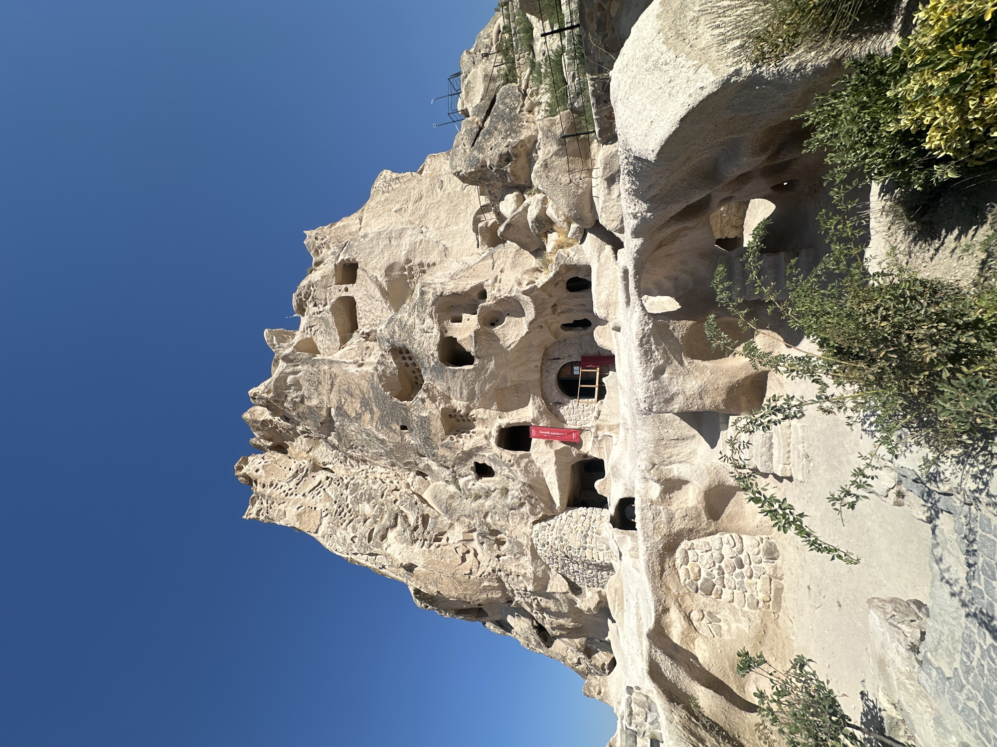 This is a low-angle, full shot of Uçhisar Castle, a prominent rock formation with numerous cave dwellings, in Uçhisar Beldesi, Turkey, under a clear, bright blue sky. The castle is a large, light-colored geological formation carved into numerous openings and hollows, suggesting a history of human habitation within the rock. Some of these openings appear to be entrances to rooms or passages. A red banner is hanging from one of the openings, featuring white text that appears to be in Turkish. Another opening nearby has wooden doors and a small window. The base of the formation is surrounded by stone pathways and low-lying greenery. Some parts of the rock face are reinforced with dry-stone masonry. The overall impression is of an ancient, sculpted dwelling within a natural rock structure.