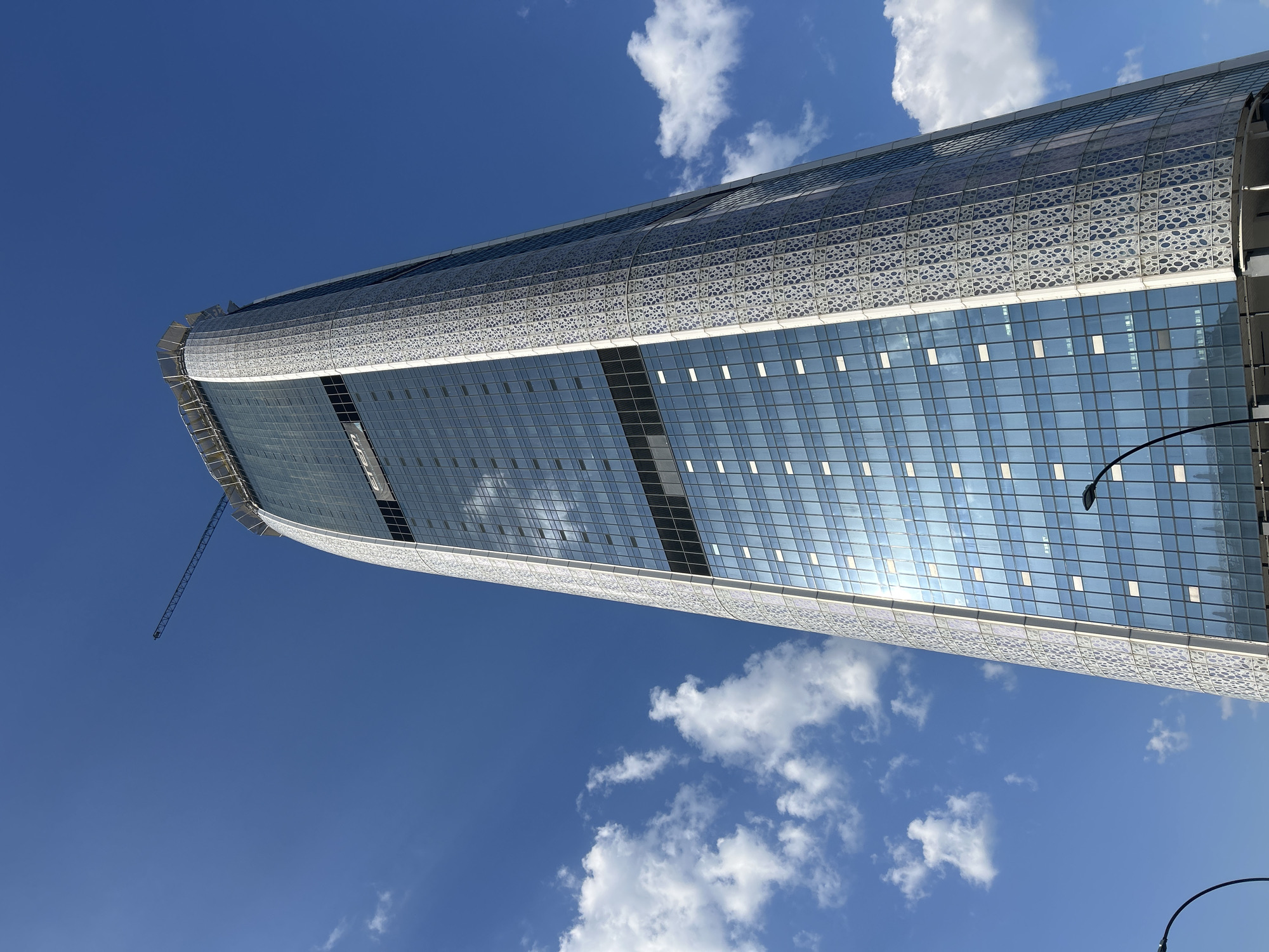 A low-angle, wide shot captures the nearly completed "Capital Fort" skyscraper under a bright blue sky with scattered white clouds. The building, located in Sofia, Bulgaria, is a modern, glass-clad structure with a distinctive patterned facade along its edges. A construction crane is visible at the very top of the skyscraper, indicating ongoing work. The sun reflects off the numerous glass panes, creating a shimmering effect. Streetlights are visible in the foreground.