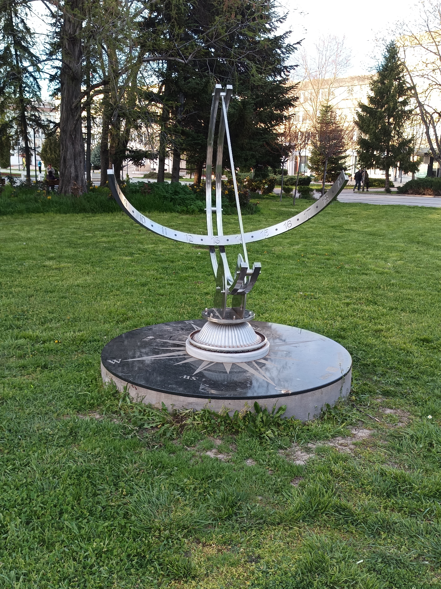 A large, modern sundial stands prominently in a green park, likely in Targovishte, Bulgaria. The sundial is constructed from shiny metal, possibly stainless steel, and features a central gnomon casting a shadow onto a curved scale. This scale displays hours marked by numbers and dots, with visible numbers ranging from 9 through 18.
The sundial is mounted on a substantial circular base made of dark, polished stone or marble. Etched onto this base is a detailed compass rose, with the cardinal and intercardinal directions clearly visible, including N, NW, W, SW, S, SE, and E. The sundial and its base are surrounded by a well-maintained lawn of lush green grass.
The background of the scene features numerous tall trees, encompassing both broadleaf deciduous trees and evergreen conifers, suggesting a mature park landscape. Through the gaps in the trees, multi-story buildings with visible windows can be seen in the distance, indicating an urban or semi-urban environment. Paved pathways wind through the park, and a few people are present: one person is seated on a park bench under a large tree on the left, and two individuals are walking along a path on the right side of the image.
The lighting suggests either early morning or late afternoon, characterized by soft, bright light and a clear sky, contributing to a peaceful and serene atmosphere. There are no specific events taking place; it's a static snapshot of a public space.