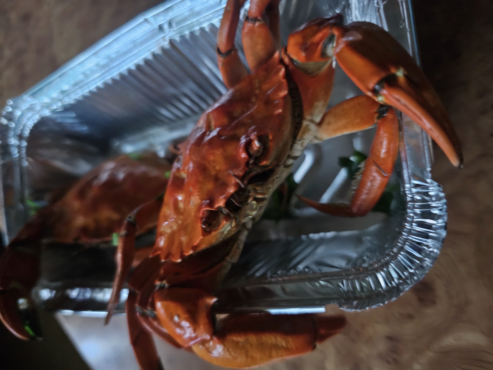 The image displays a close-up, low-angle shot of two cooked crabs arranged in a foil container. The crabs are bright orange and appear to be whole, with visible claws and legs. The foil container has a textured, rippled interior and a blue-tinged rim. The crabs are positioned diagonally within the container, with the larger crab in the foreground and a portion of a second crab visible behind it. The background is out of focus, showing a wooden surface. The lighting is dim, with some reflections visible on the crabs' shells and the foil. There are no people, actions, or interactions depicted in the image. The scene does not provide any visual information to place it in Skadovsk, Ukraine.