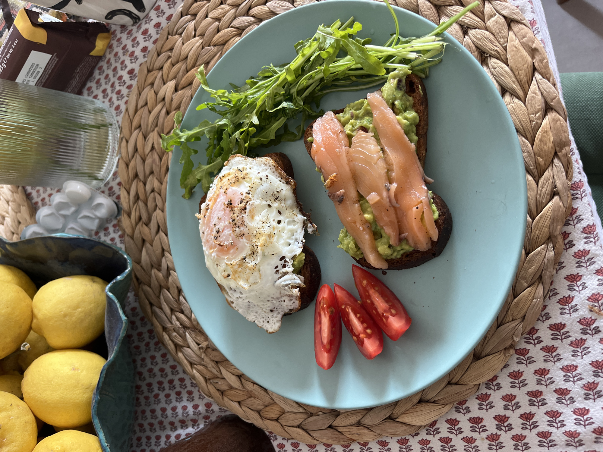 The image displays a vibrant, healthy meal, likely breakfast or brunch, served on a light blue round plate. On the plate are two slices of dark-colored toast. One slice is topped with mashed avocado and several delicate strips of smoked salmon. The other toast features mashed avocado beneath a fried egg, seasoned with black pepper, its yolk still slightly runny. A generous portion of fresh green arugula leaves and three bright red tomato wedges complete the meal.
The plate rests on a round woven placemat made of natural, light-brown fibers, which in turn lies on a white tablecloth adorned with a repetitive pattern of small red floral motifs. To the left of the plate, a clear ribbed glass and a decorative white ceramic object are visible. In the bottom left corner, a dark blue or teal ceramic bowl with an undulating rim is brimming with bright yellow lemons. A dark-colored food package is partially visible at the top left, displaying legible text including "NET WT. 4.4 OZ", "PD 12/08/2020", and "EX 12/08/2022", along with "L 0000 2" and "25g e". A glimpse of green upholstered furniture is seen on the far right, suggesting a comfortable indoor setting. The bright, natural lighting indicates the picture was taken during the daytime in Ramat Gan, Israel.