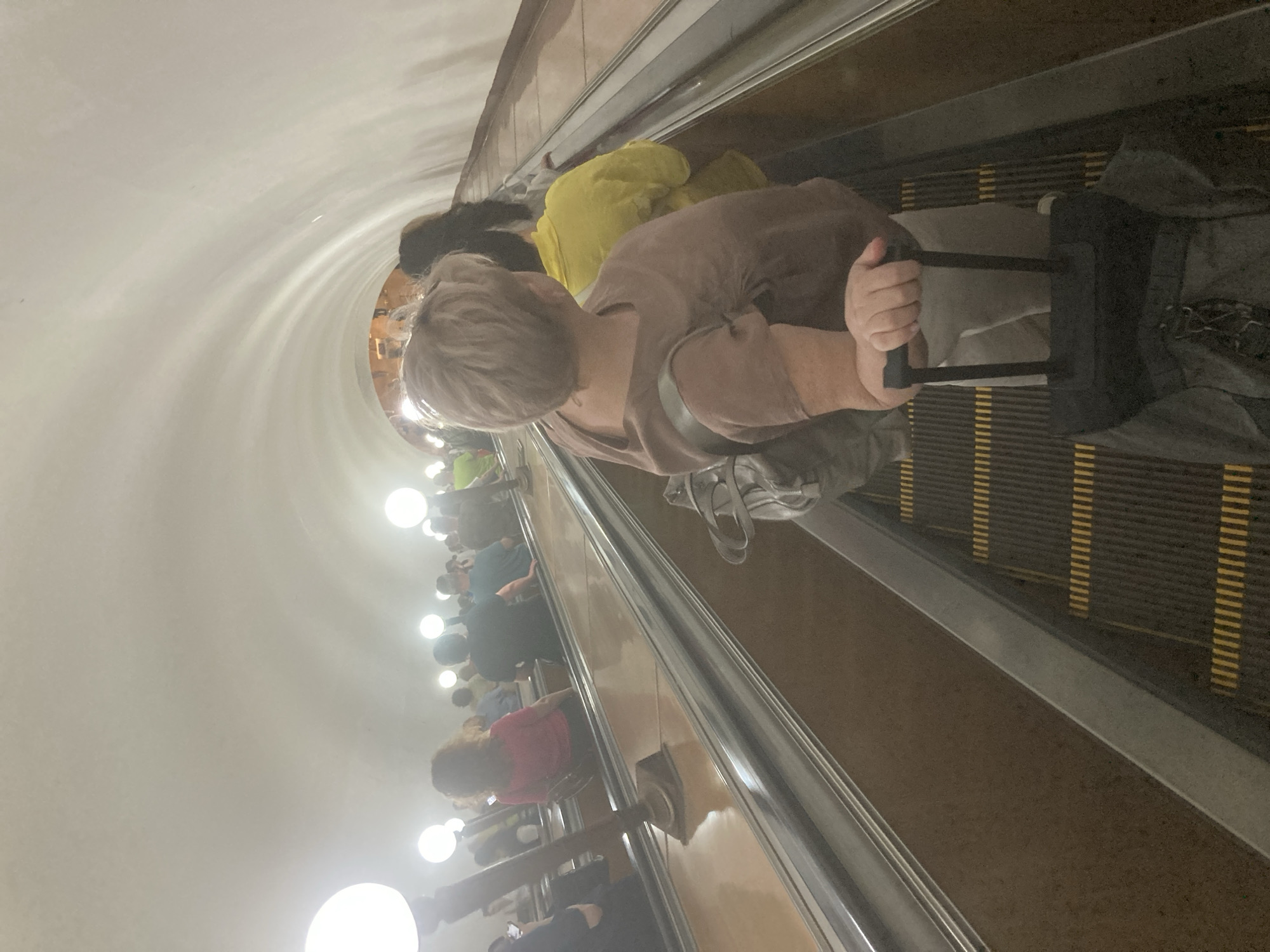 The image captures a scene inside a Moscow Metro station in Russia. An escalator is visible, carrying passengers upwards. An adult female, likely in her sixties based on her gray hair, is riding the escalator with a dark-colored suitcase in her right hand. She is wearing a light brown shirt and light-colored pants. The metro station is illuminated by lights. The ceiling has a curved design. Other passengers are seen on the escalator behind the main subject, including people wearing yellow and pink clothing.