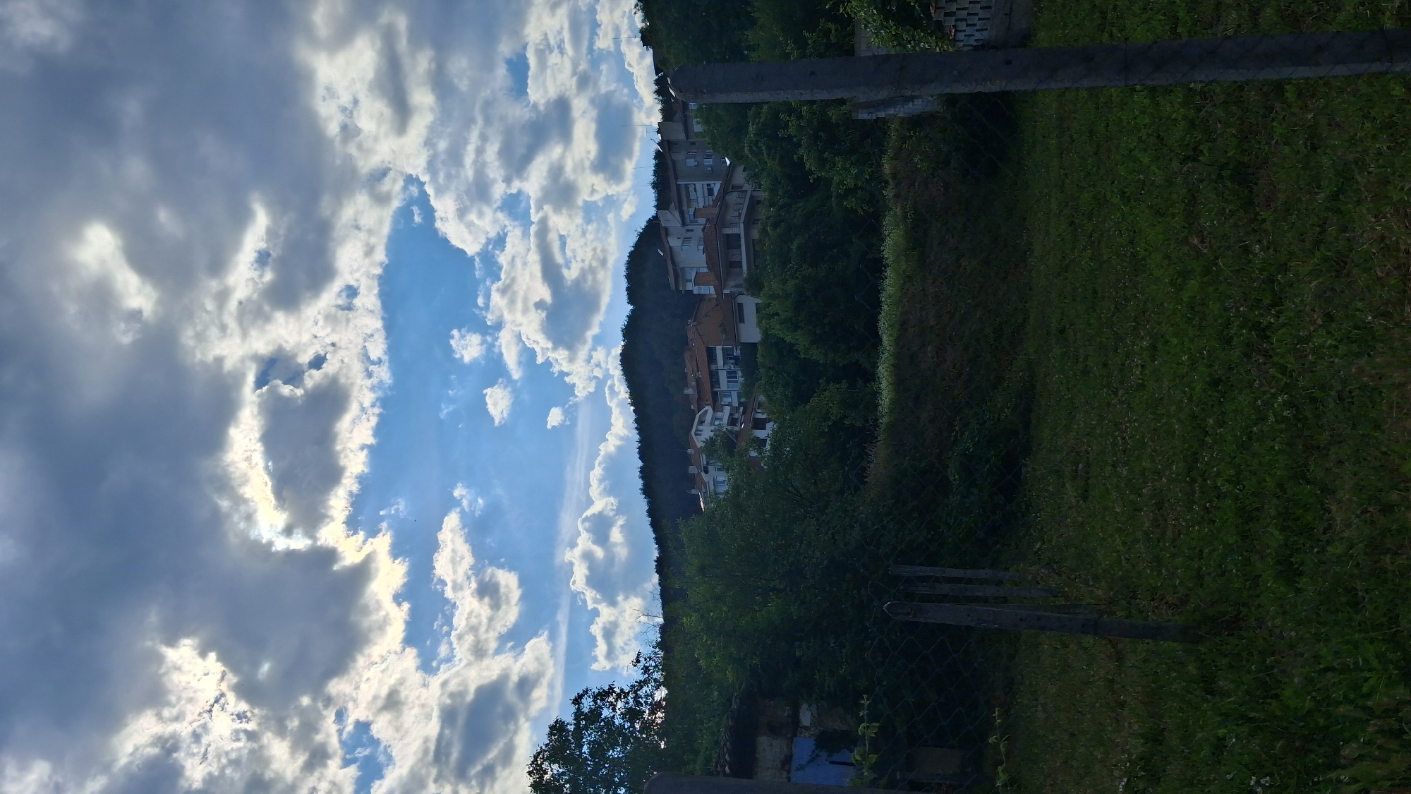 The image shows a view of buildings on a hillside under a cloudy sky. The sky is a mix of blue patches and dramatic grey and white clouds, with sunlight breaking through in some areas. Below the sky, a green, tree-covered hillside is visible, with several buildings clustered on it. These buildings appear to be residential, with a mix of architectural styles, including tiled roofs and multi-story structures. In the foreground, there is a grassy area with a wire fence and concrete posts. The overall impression is of a somewhat overcast but potentially sunny day in a residential area on the outskirts of a town. Given the context, this could be a scene in Gotse Delchev, Bulgaria, depicting a typical residential neighborhood situated on the slopes surrounding the city. No people are visible in the image.