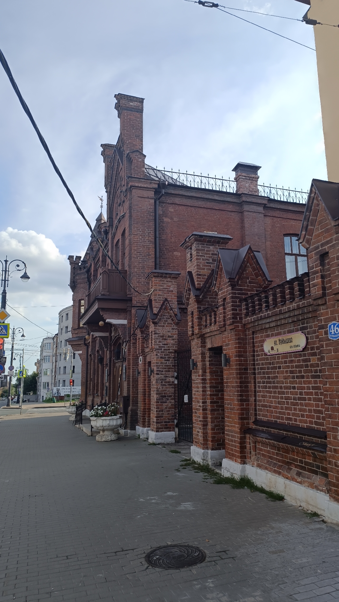 The image depicts the exterior of a historical brick building, identified as located at 46 Kuybyshev Street in Novokuybyshevsk, Russia. The building features intricate brickwork, decorative chimneys, and a wrought-iron fence. A street sign indicates the address "ул. Куйбышева 46" (Kuybyshev Street 46). The building is situated on a paved sidewalk adjacent to a street. In the background, other buildings and street infrastructure, including traffic lights and street lamps, are visible. The sky is partially cloudy. There are no people present in the image.