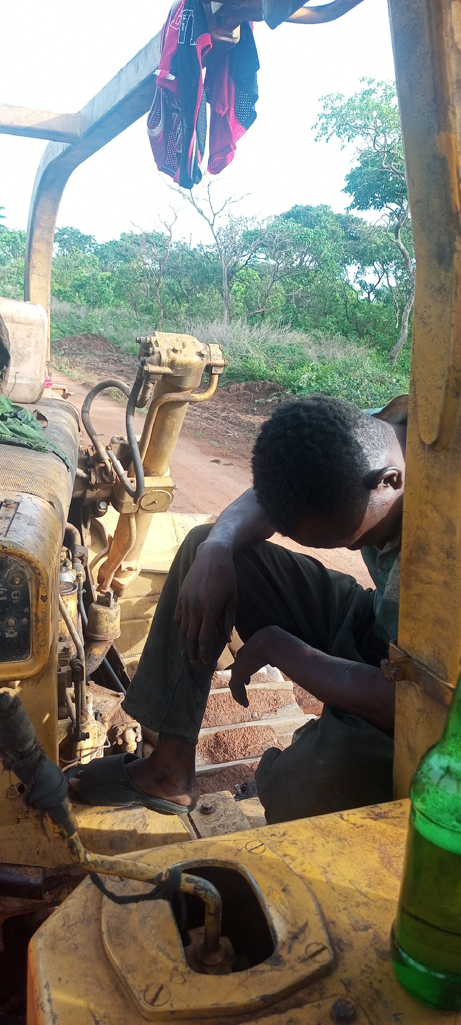 A young man is resting inside the cabin of a large, yellow piece of heavy machinery, likely a bulldozer or similar construction vehicle. He is seated on the right side of the frame, leaning forward with his head down, appearing to be asleep or deeply contemplative. His dark, curly hair is visible, and his skin is dark. He wears dark pants and black flip-flops. His hands and arms show signs of dirt or grime, suggesting manual work.
The machinery itself is old and robust, painted yellow, with various levers, pipes, and gauges visible on the console to the left of the man. Faint labels like "OFF", "WASH", and "RESET" can be partially discerned on some controls. Above the man's head, two pieces of red and black athletic-style clothing, possibly jerseys, are hung to dry or air out from an overhead bar of the vehicle's frame. To the far right in the foreground, a green glass bottle is visible, resting on a part of the machinery.
The setting is outdoors, under bright daylight, indicated by the strong shadows and clear sky. In the background, a dirt road or path stretches into the distance, flanked by lush green trees and bushes, suggesting a rural or construction site environment. The ground visible behind the man and the machinery is reddish-brown, typical of laterite soil. The overall impression is one of a break from labor in a warm, outdoor environment in Otunade, Nigeria.
