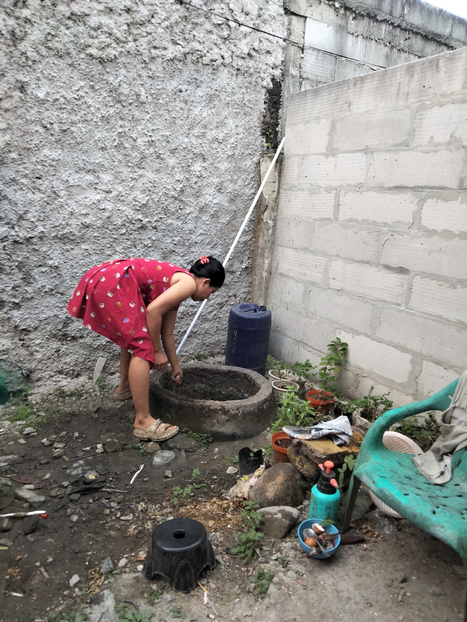 In Cariu, Indonesia, a young woman in a red polka-dot dress is bending over a circular concrete basin, appearing to wash something by hand. She has her hair tied back with a red clip. The scene is set in a cluttered outdoor area with rough, textured walls made of concrete blocks and plaster. Various objects are scattered around: a large blue plastic barrel, several potted plants, rocks, a turquoise spray bottle, a small blue bowl containing eggshells and other debris, and a green plastic chair with clothing draped over it. The ground is a mix of dirt, gravel, and debris, with some patchy green vegetation growing. A white PVC pipe runs diagonally up the textured wall.