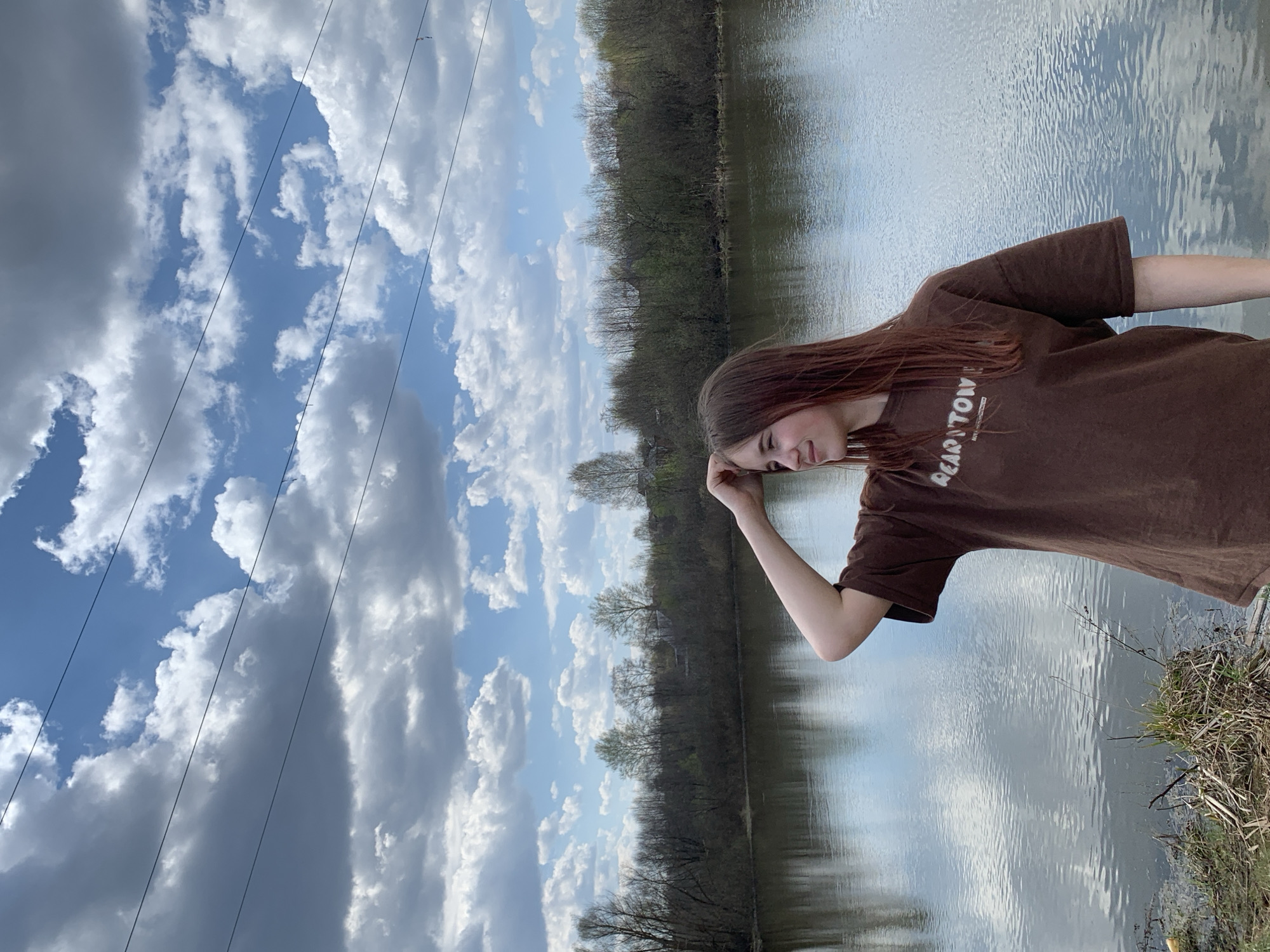 A young woman with long, reddish-brown hair stands near a body of water. She is wearing a brown t-shirt. The background consists of a lake, trees, and a cloudy sky with power lines visible. The location is likely near a natural area, but the specific city of Фоки, Russia, cannot be confirmed from the image alone.