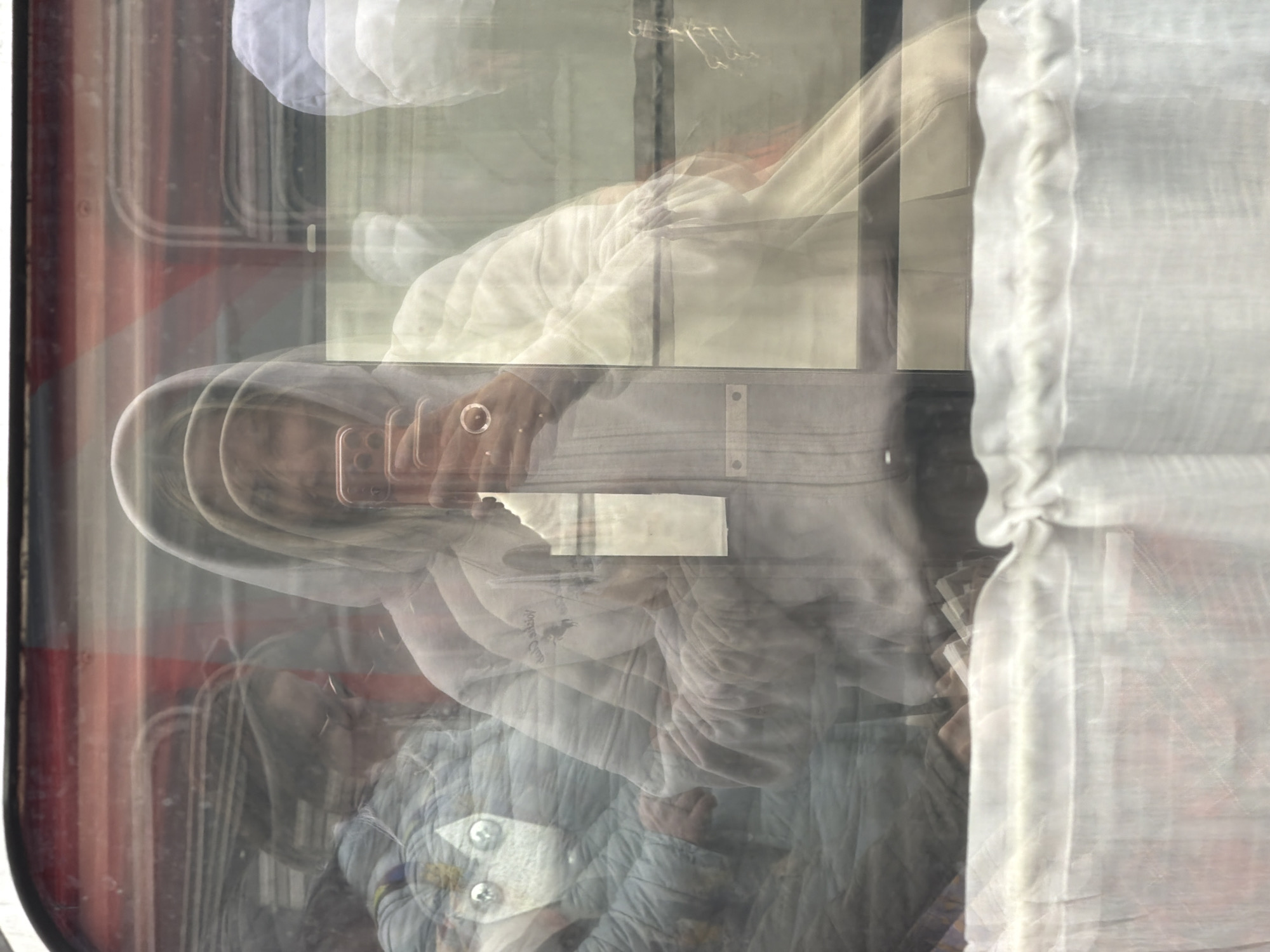 The image appears to be taken through a window, likely of a train or other public transportation in Moscow, Russia. The reflection on the glass creates a layered and slightly blurred effect. In the reflection, a person is visible, holding a mobile phone up, possibly taking a photograph. The person is wearing a light-colored hooded jacket. The interior of the train or bus is also reflected, with some details of seats and a white curtain visible. The red exterior frame of the window is also visible, suggesting it's a train or bus window. Due to the reflections and blurry nature, specific details are hard to discern.