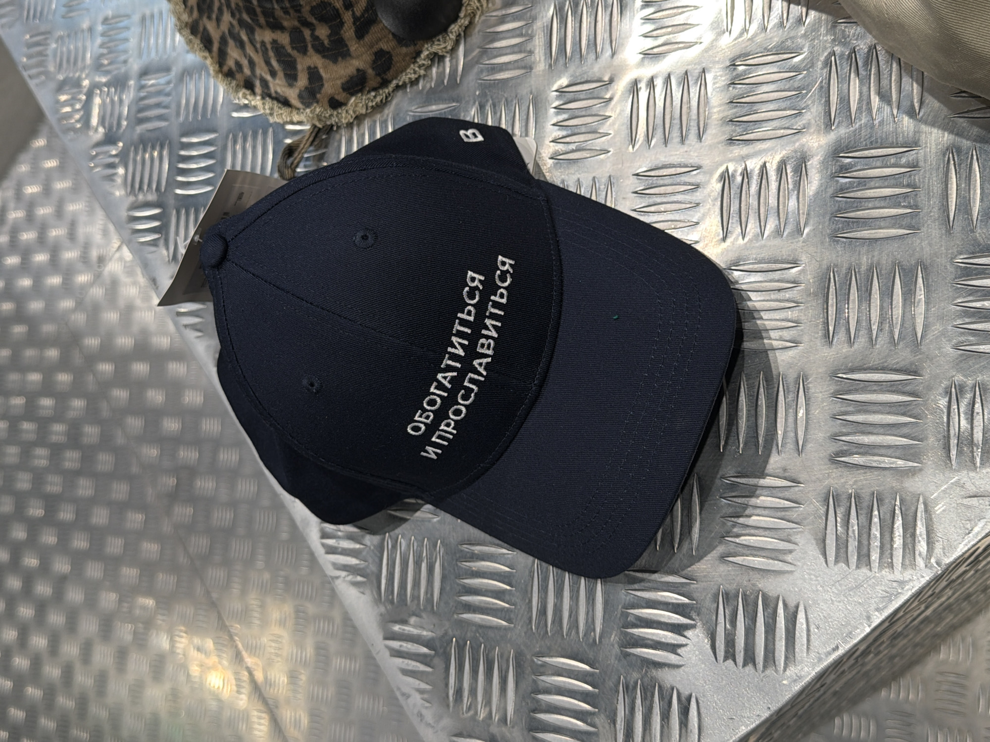 A dark blue baseball cap with white Russian text embroidered on it is placed on a metallic surface with a diamond plate pattern. The text on the cap reads "ОБОГАТИТЬСЯ И ПРОСЛАВИТЬСЯ" which translates to "Get Rich and Glorified". A small white letter "B" is visible on the side of the cap. Next to the cap, a portion of a leopard-print fabric is visible, possibly from another item of clothing or accessory. The metallic surface suggests an indoor environment, possibly a store or an exhibition space. There is no indication of the specific location within Moscow, Russia. The image is a close-up, top-down view of the cap and the surrounding surface.