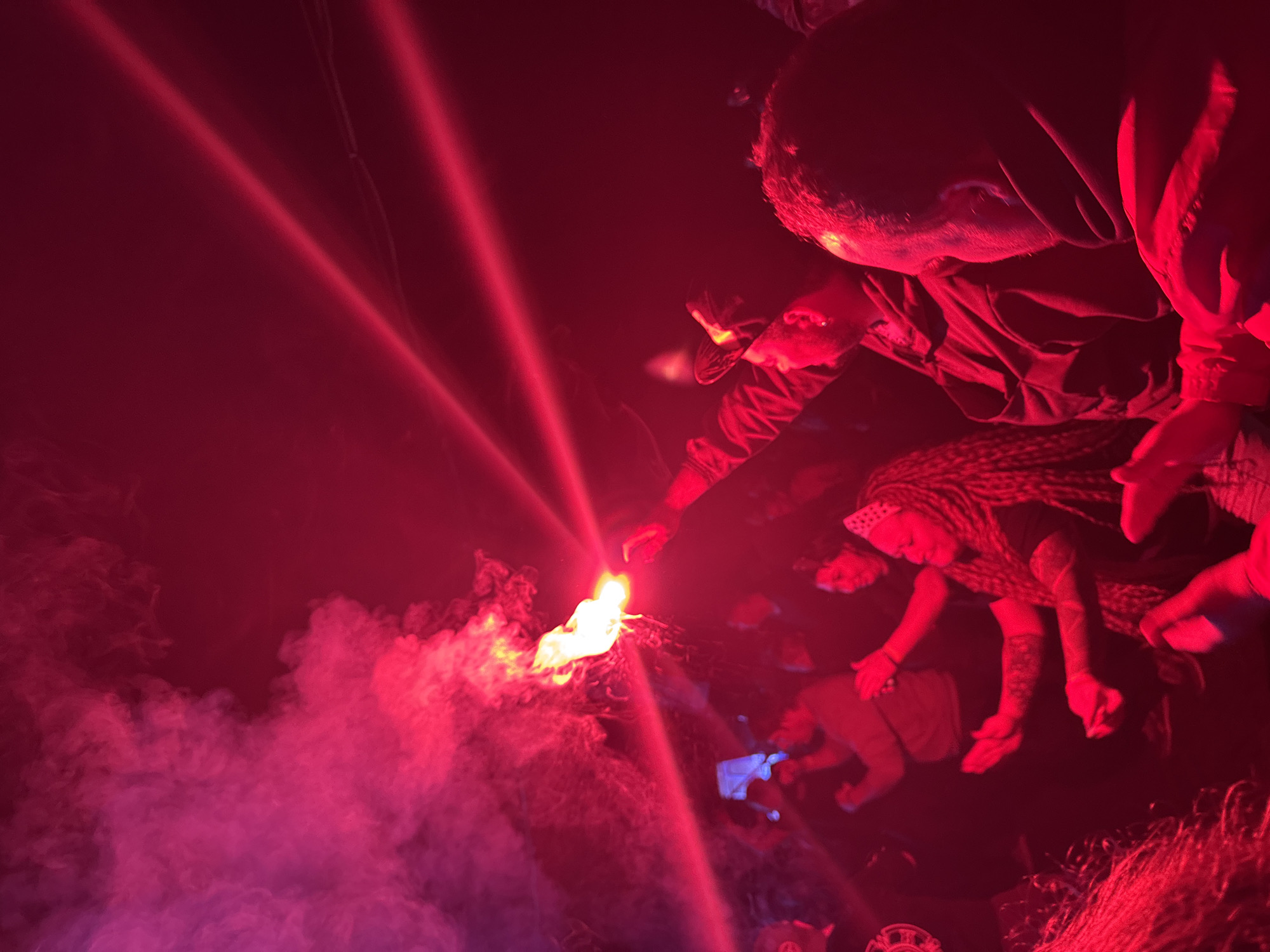An indoor gathering in Большая Кивара, Russia, is depicted under intense red artificial lighting and significant atmospheric smoke. In the central-left, an individual holds a burning orange-yellow flare, which emits a bright flame, sparks, and dense smoke that diffuses into the environment. Two distinct, diagonal red laser beams traverse the upper right portion of the frame. Numerous people are present, largely obscured by the low light and red illumination. In the mid-right, a woman with long, braided hair and visible arm tattoos is captured with her arms raised, suggesting movement or dancing. Other figures throughout the scene display similar gestures, with some individuals in the left-midground seen holding mobile phones, one of which has a glowing blue-white screen, possibly recording the event. The scene conveys a dynamic social gathering or performance.