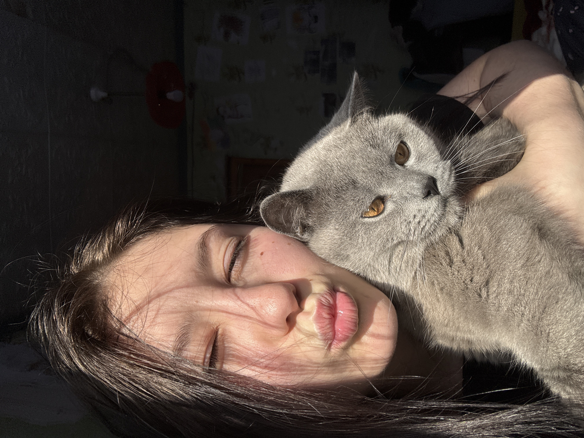 A young woman with dark hair and closed eyes lies on her back, her face puckered into a kiss. A gray cat with amber eyes is nestled against her neck and upper arm. Sunlight falls across the woman's face and the cat's fur, while the background is darker, revealing a partially visible wall with pictures and a red lamp. The location is not evident from the image and cannot be determined to be Фоки, Russia.