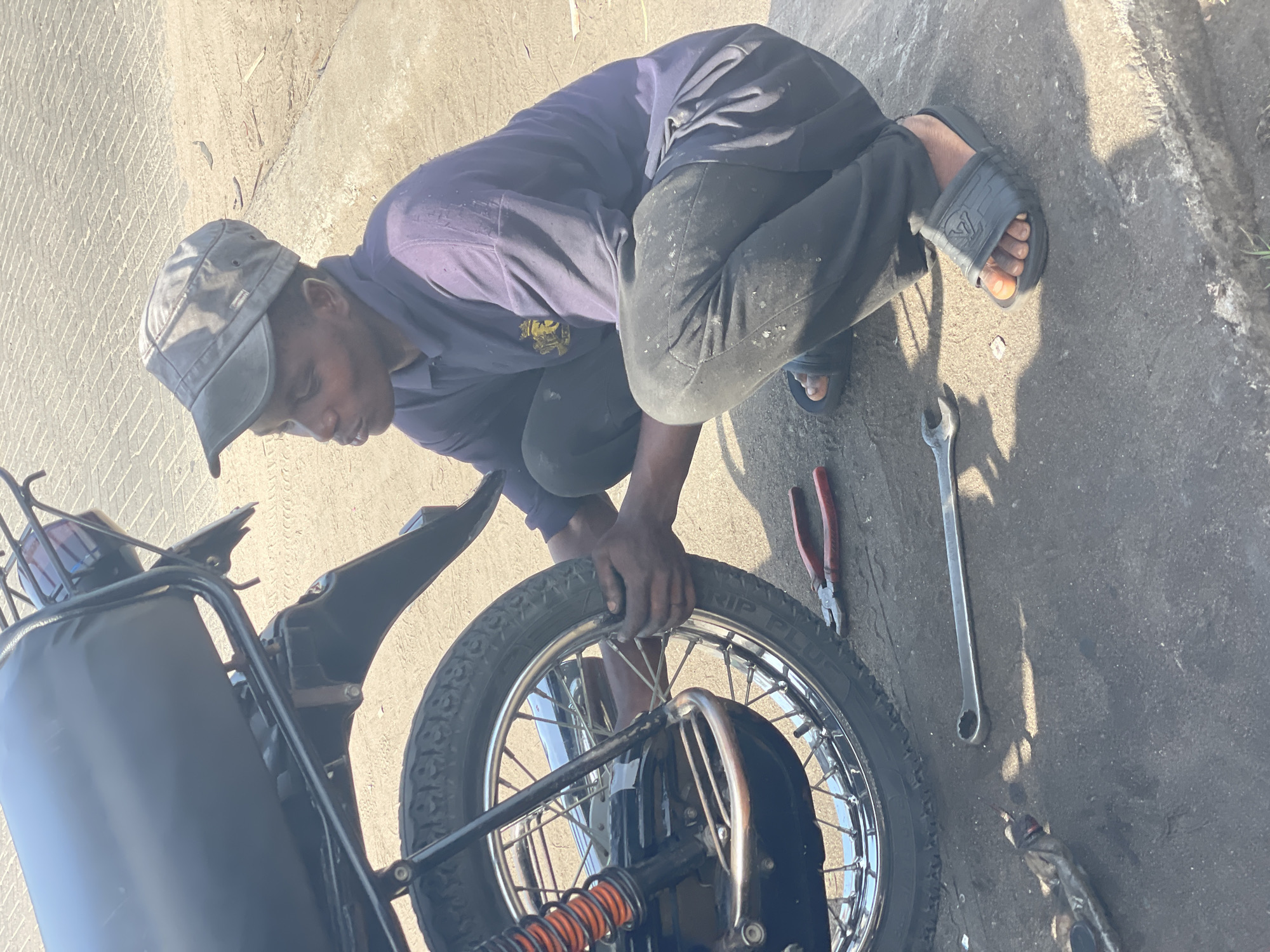A man is crouched next to a motorcycle, working on its rear wheel. He is wearing a grey cap, a blue-purple long-sleeved shirt, and dark pants. He is also wearing dark-colored sandals. Next to the wheel, there are pliers and a wrench lying on the ground. The location is implied to be Badagry, Nigeria, based on the user's input.
