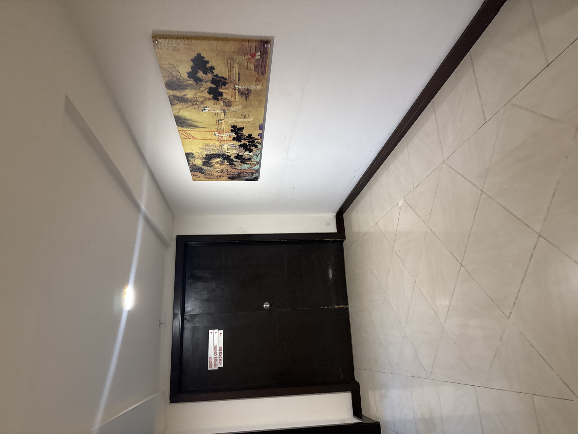 The image depicts a hallway with a double dark wooden door on the left. Above the door, three white signs with red text indicate "DOCTOR," "FITNESS CENTER," and "HAIR DRESSER," each with an arrow pointing to the right. The hallway floor is tiled with large beige squares arranged in a diamond pattern. On the right wall, a rectangular painting with an East Asian motif hangs. The painting features figures, trees, and a bridge against a golden background. The walls are white, and a dark brown trim runs along the base of the walls. A light fixture is visible on the ceiling, casting a bright light. The environment appears to be an indoor corridor within a building.
