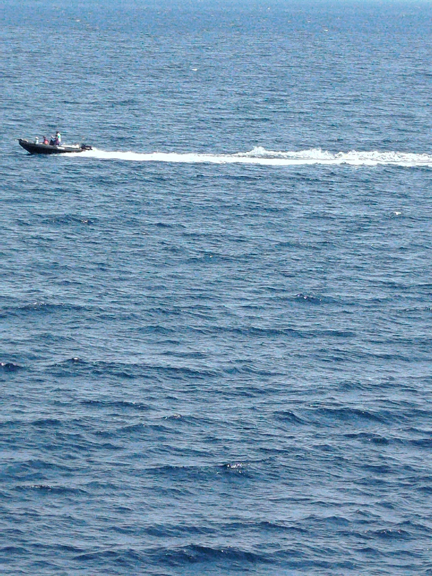 A small, dark-colored motorboat with three occupants is moving at speed on a body of water, leaving a white wake. The boat is positioned in the upper left quadrant of the frame. The water is a deep blue with moderate wave action. The sky is a lighter blue and appears to be slightly hazy. The scene is devoid of any landmarks or identifiable city structures, making it impossible to determine its specific location within Sithonia Municipal Unit, Greece, or any other city.