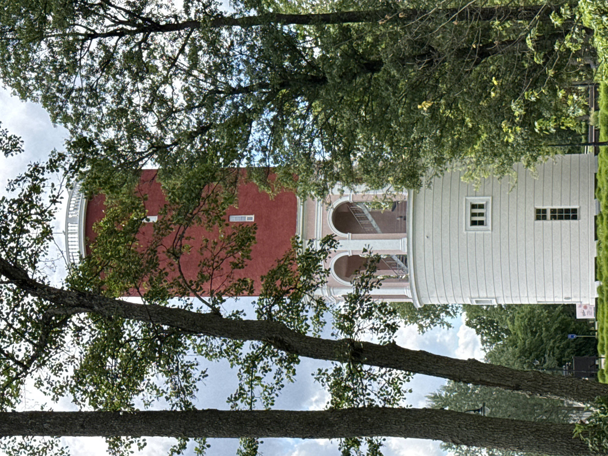 A tall, cylindrical water tower with a red upper section and a white lower section is partially obscured by trees in Jūrmala, Latvia. The tower has white arched openings with visible stairs behind them on the pinkish-white middle section. The white lower section features horizontal grooving and several rectangular windows. A white railing encircles the top of the red section. The surrounding environment consists of lush green trees with dark trunks and dense foliage, and a patch of well-maintained green grass in the foreground. The sky above is partly cloudy. There are no people or animals visible in the image.
