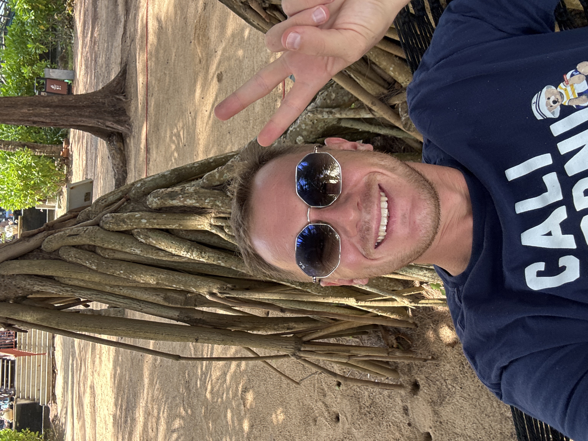 A man is taking a selfie in a hammock, possibly at Bang Niang Beach in Ban Khuk Khak, Thailand. He is wearing sunglasses and a navy blue t-shirt with graphics on it. He is smiling and holding up two fingers in a peace sign. The background shows sand, large tree roots, and lush green vegetation. In the distance, there are stairs and a trash can. The man appears relaxed and is enjoying the tropical environment.
