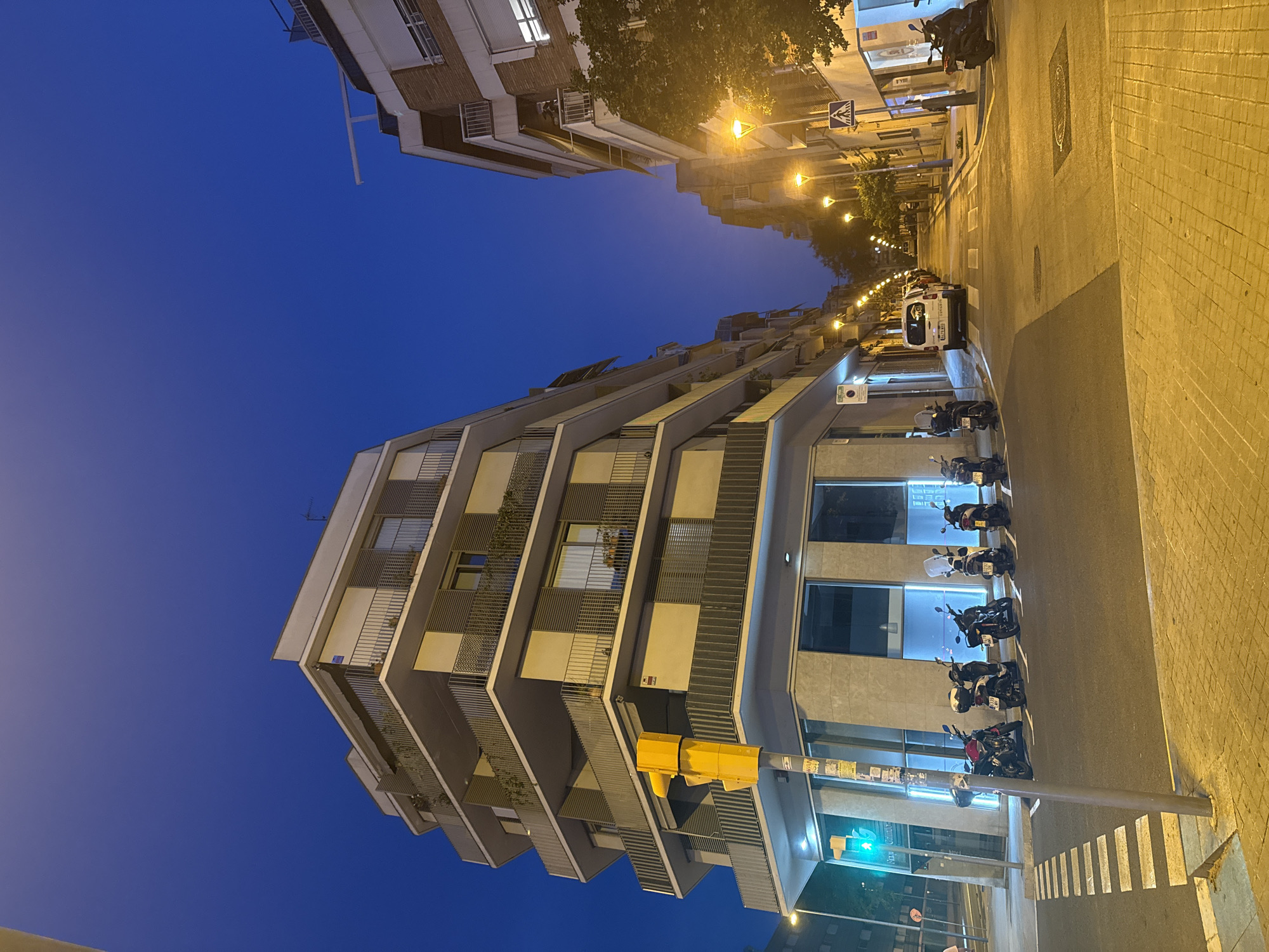 This is a photograph taken at night in Barcelona, Spain, on a street with multi-story buildings on either side. The sky is a deep twilight blue. On the left, a modern, angular building with multiple balconies and horizontal louvers is prominently featured. A yellow traffic signal is visible at the corner of this building, with a green light illuminated.
In front of the building on the left, several parked scooters are lined up. The street is paved with asphalt, and there is a pedestrian crossing with white stripes. The sidewalk is paved with yellow bricks.
The street recedes into the distance, lined with streetlights casting a warm, orange glow. More buildings are visible along the street, some with balconies and awnings. A white van is parked on the right side of the street. A street sign with a pedestrian symbol is also visible on the right. The overall impression is an urban street scene at dusk or early night, with artificial lighting creating distinct shadows and highlighting the textures of the urban environment. No people are directly visible in the frame, suggesting a quiet moment in the city.
