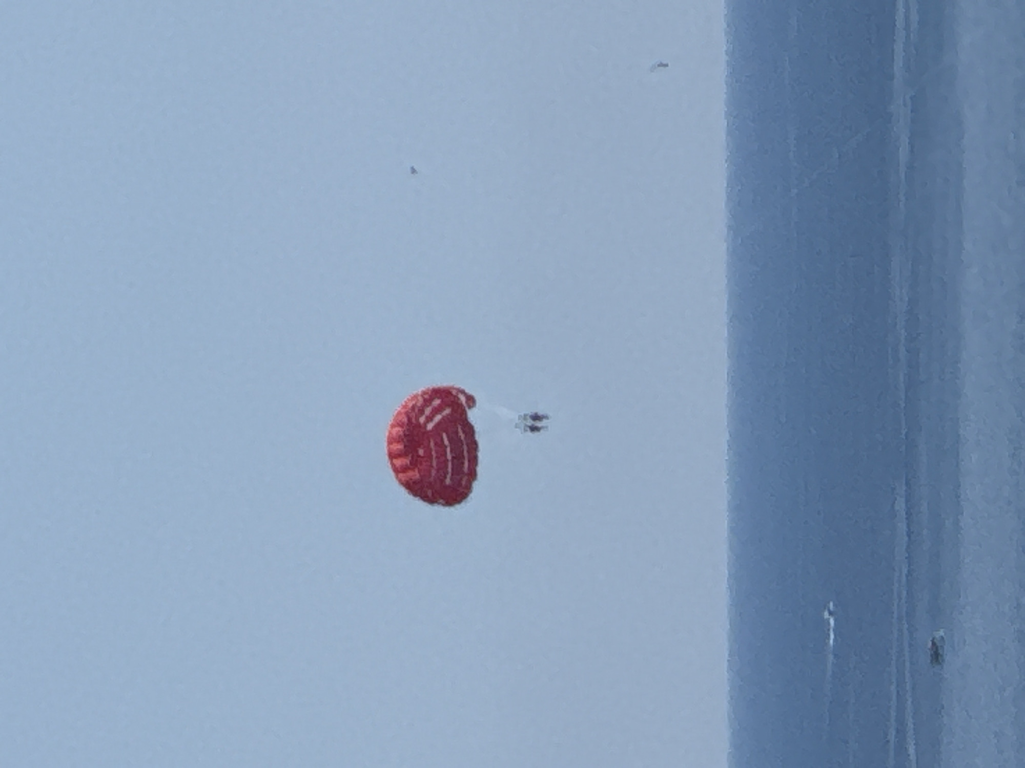 In the sky over the sea, a red and white parachute is visible with two people harnessed below it, parasailing. The sea is calm and blue with a distant shoreline. The sky is a pale blue and hazy.