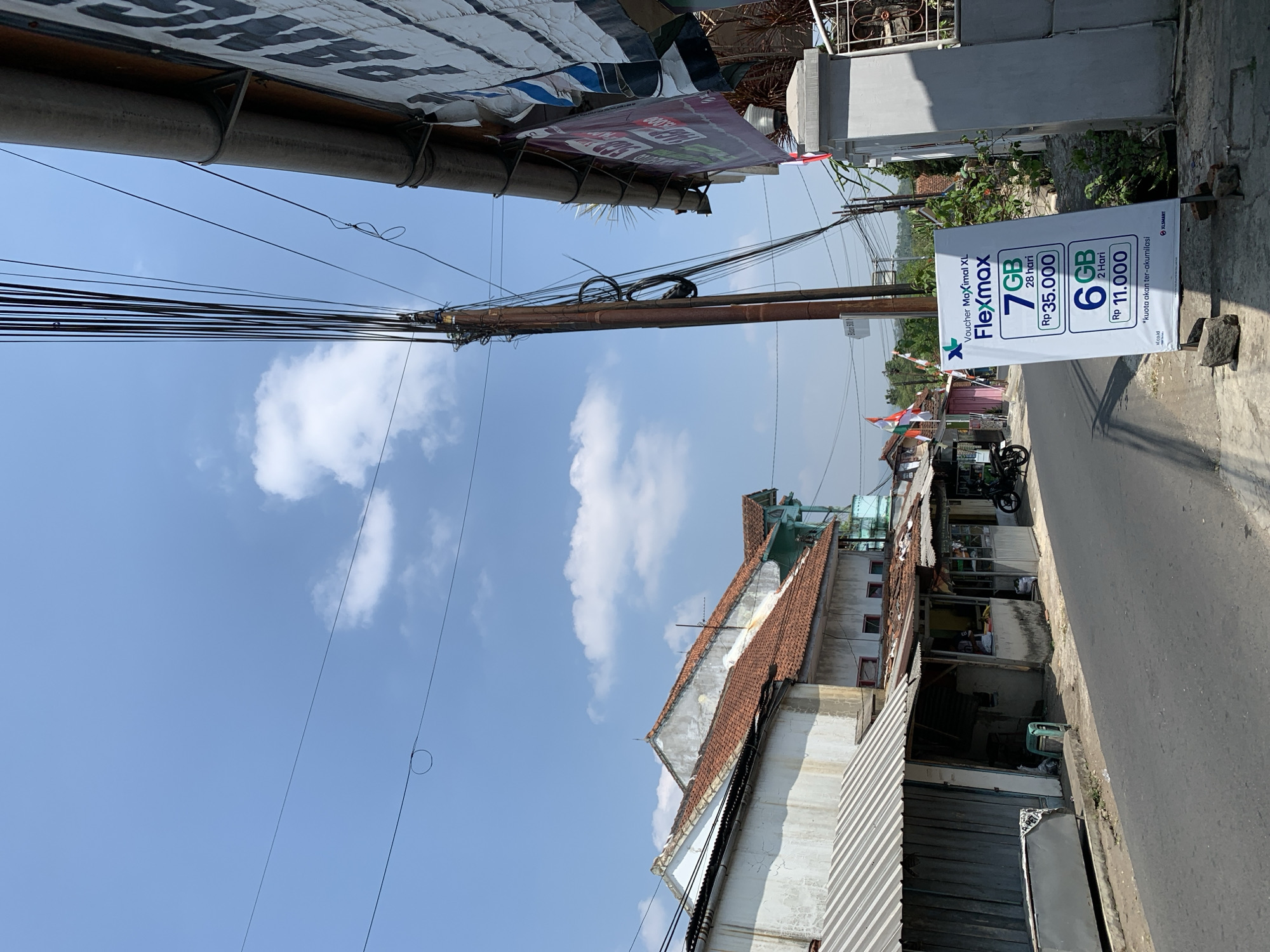 A street scene in Ciamis, Indonesia, features a roadside advertisement for XL Axiata's Flexmax data packages. The advertisement displays two options: 7GB for 28 days at Rp 35,000 and 6GB for 2 days at Rp 11,000. The scene includes a road, buildings with red-tiled roofs, utility poles with numerous wires, and a clear blue sky with scattered clouds. A motorcycle is parked near one of the buildings. Several Indonesian flags are visible, suggesting a national holiday or patriotic display. The overall impression is of a typical urban environment in a tropical region.