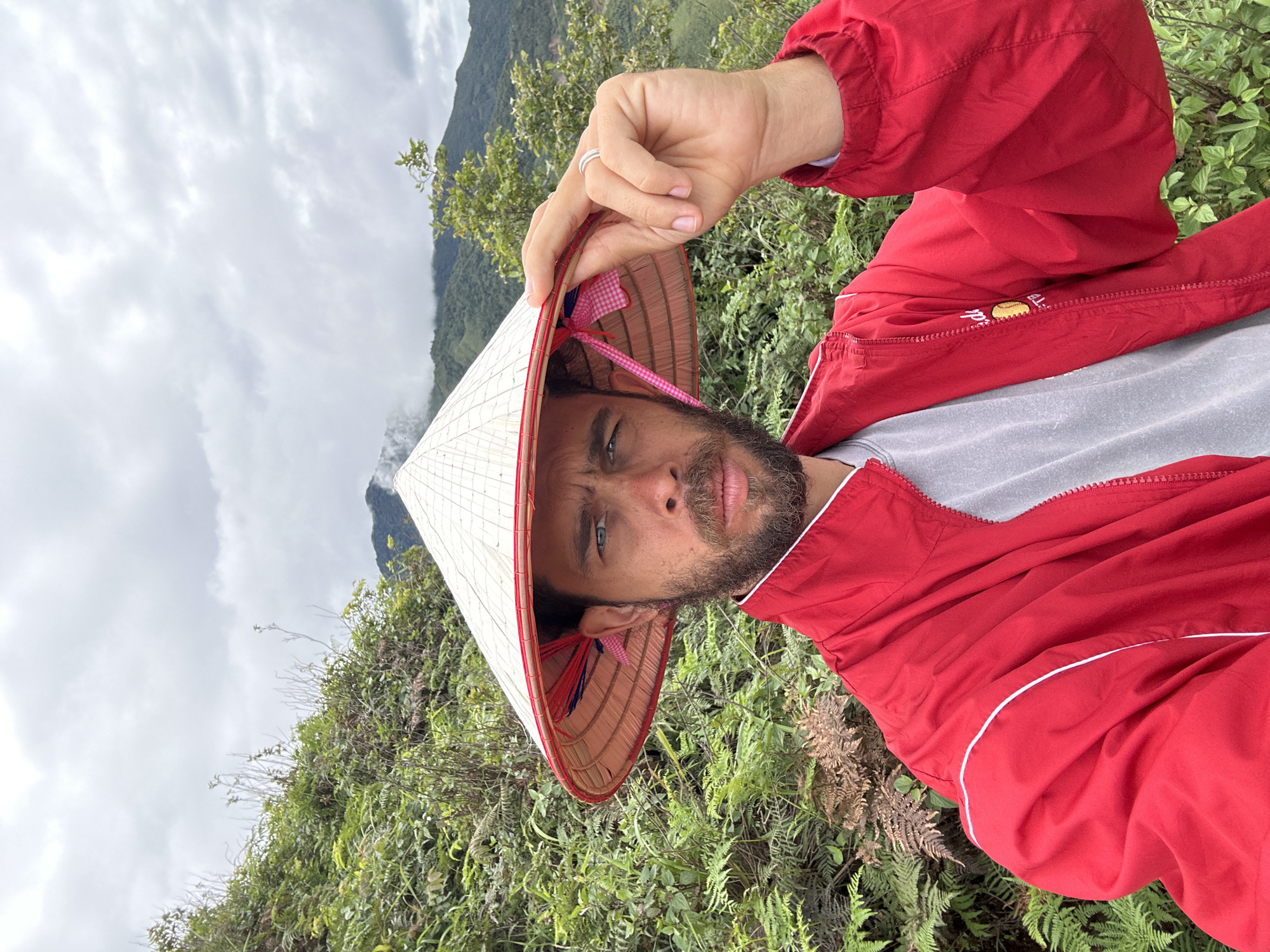 In Ta Xua, Vietnam, a man wearing a conical hat and a red jacket is taking a selfie. The man has a beard and is looking at the camera with a serious expression. He is holding the brim of his hat with his right hand, revealing a ring on his finger. The background is a lush, green landscape with mountains in the distance. The sky is overcast, and there is a slight mist rising from the mountains. The man is standing on a slope covered in ferns and other vegetation.