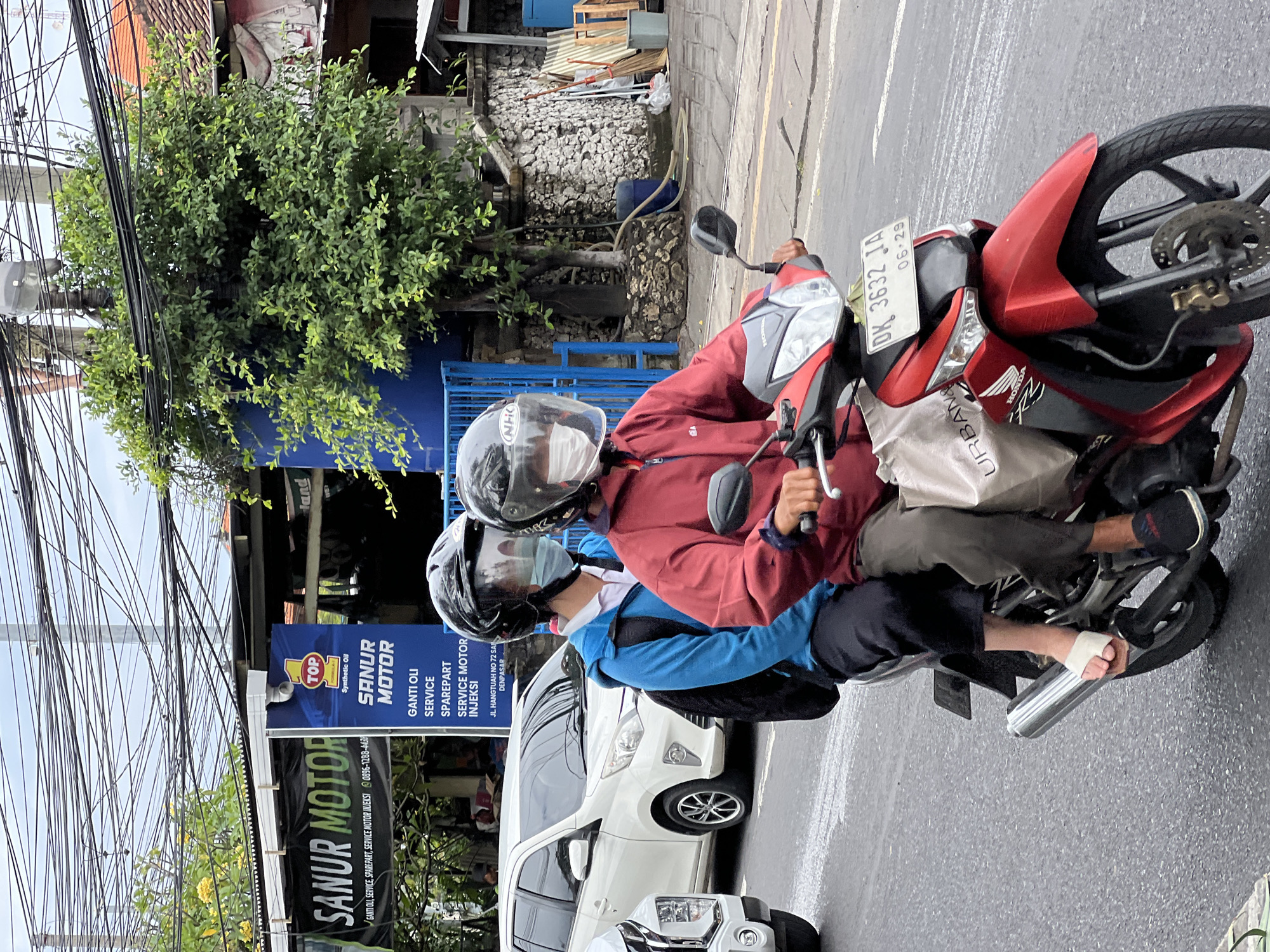 Two people are riding a red and black motorcycle in Denpasar, Indonesia. The person in front, the driver, is wearing a red jacket and a black helmet with a clear face shield. They are wearing dark pants and black sneakers. The person in the back is wearing a blue jacket and a black helmet with a clear face shield. They are wearing dark pants and white flip-flops. Both individuals are wearing face masks. A white tote bag with the word "URBAN &" is hanging from the front of the motorcycle. The motorcycle has a license plate that reads "DK 3632 JA" and "06-29". In the background, there is a white car, a sign that says "SANUR MOTOR", and a tree. The sky is overcast.