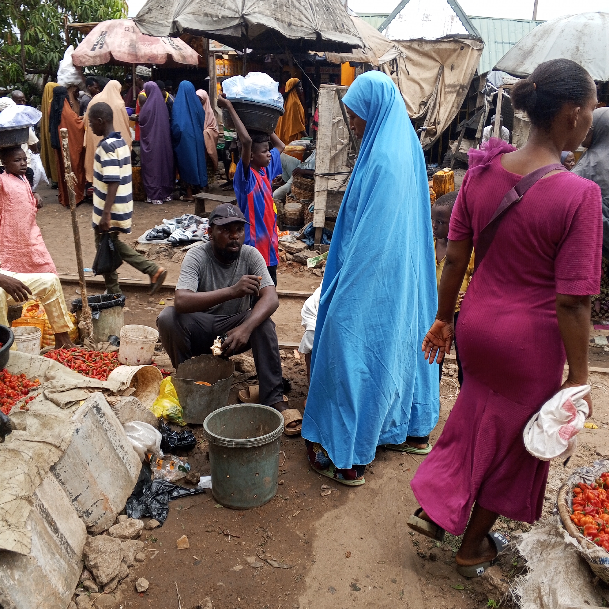 The image depicts a bustling outdoor market scene in Maiburiji, Nigeria, during what appears to be an overcast day. The ground is unpaved, covered in dirt, and strewn with debris like plastic bags, rocks, and remnants of fabric.
Numerous people are visible, moving through and congregating within the market. In the foreground, a man sits on the ground, dressed in a grey t-shirt and a black baseball cap, with a beard. He is surrounded by buckets and a substantial pile of bright red peppers, indicative of a vendor selling produce. To his right, a woman stands prominently in a vibrant blue hijab or veil, looking towards the activity within the market. To her right, another woman, seen from behind, walks away, wearing a magenta-colored dress with a shoulder strap and holding a white cloth in her hand. A child's face is partially visible behind the blue-veiled woman.
Further into the scene, a young boy in a blue and red striped shirt skillfully carries a large black basin filled with plastic bags, possibly containing water or ice, on his head. Another boy in a striped shirt walks in the mid-ground. The background is crowded with more market-goers and makeshift stalls covered by tarpaulins and large, functional umbrellas, one of which clearly displays the number "01." The overall impression is one of a vibrant, active local market where daily commerce and community interaction occur amidst a somewhat raw and utilitarian setting.