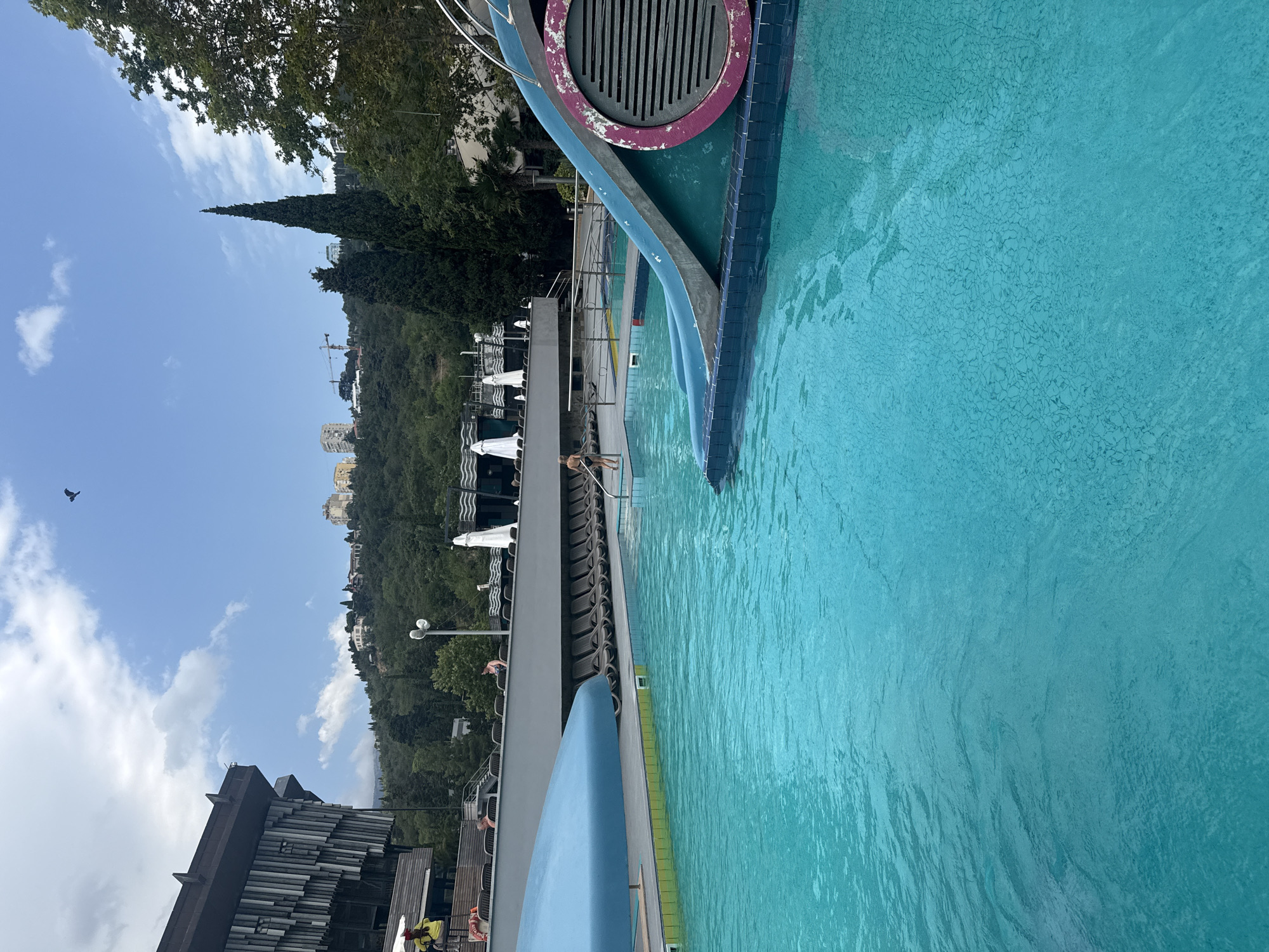 The image depicts an outdoor swimming pool area with a bright blue pool. A blue slide is positioned at the edge of the pool, partially submerged. In the background, there is a building with a modern, angled facade and several rows of lounge chairs, some with umbrellas. A person in a black bikini is standing at the edge of the pool near the slide. Further back, a lifeguard in a yellow shirt and red shorts sits on a chair. Beyond the pool area, there is a hillside covered in trees and buildings, with a clear blue sky above. A bird is visible flying in the sky. The location is identified as Yalta, Ukraine.